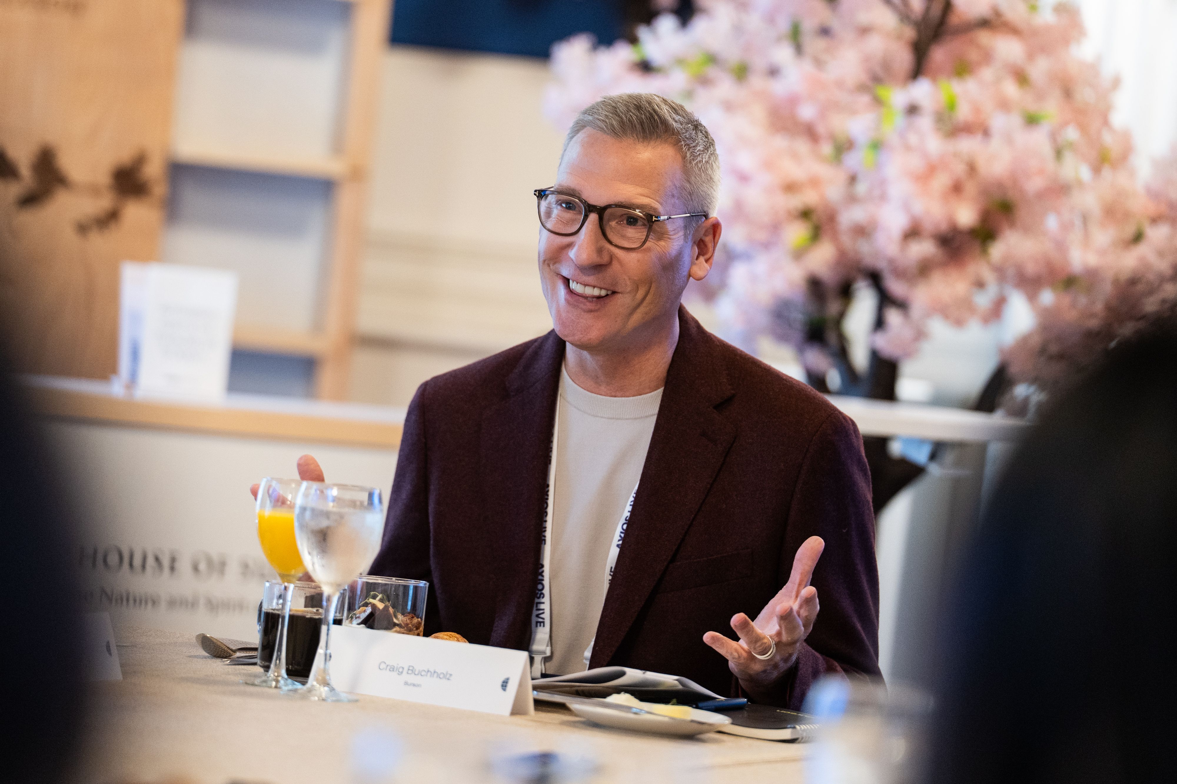 Smiling man with gray hair and glasses in a burgundy blazer sits at a table with drinks; a name card reads "Craig Buchholz." A pink floral backdrop and blurred foreground frame the scene as he gestures during a discussion.