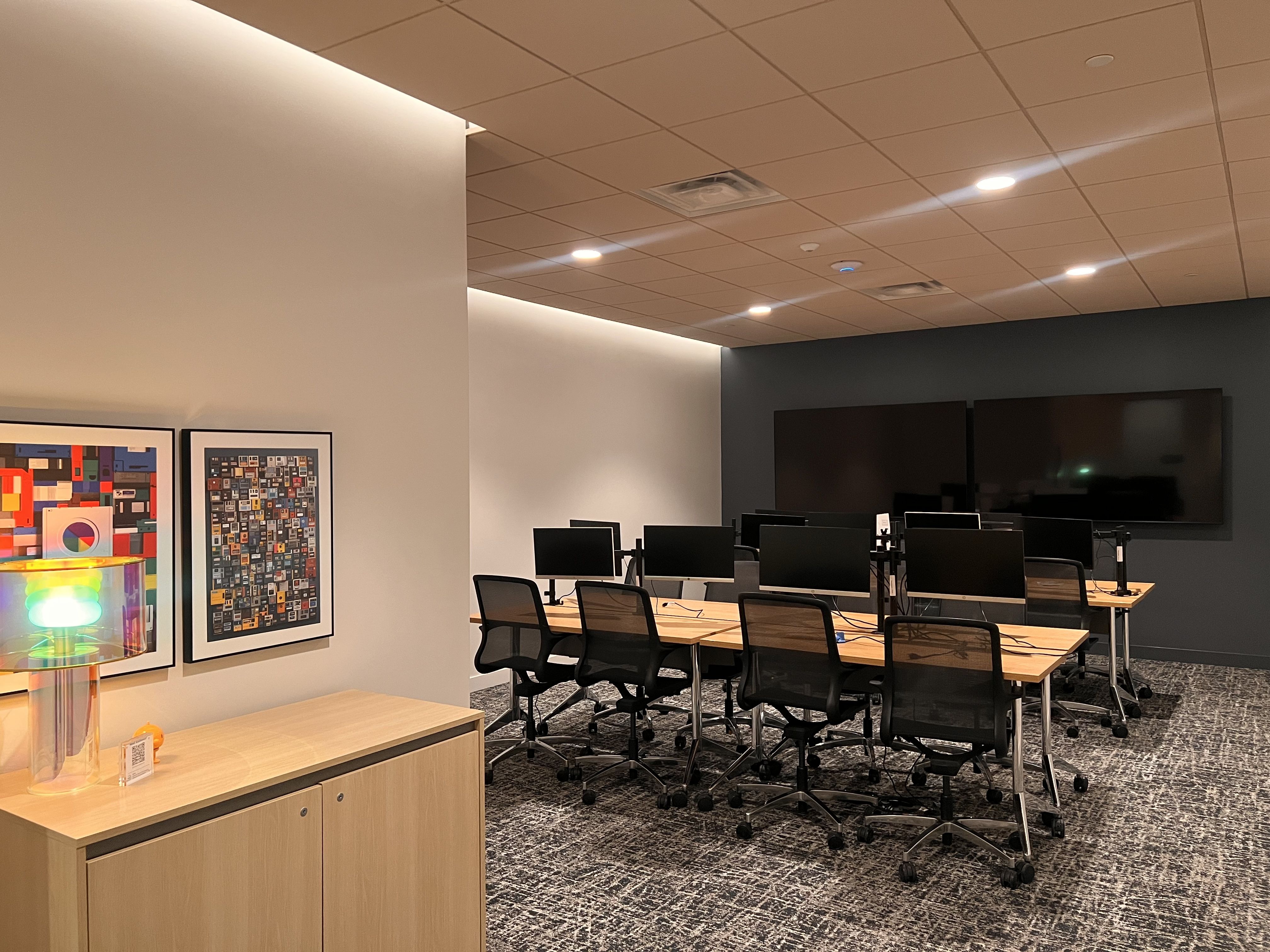 Modern office meeting room with wooden desks, black mesh chairs, multiple computer monitors, two large wall-mounted screens, colorful artwork, and a translucent decorative lamp on a beige cabinet.