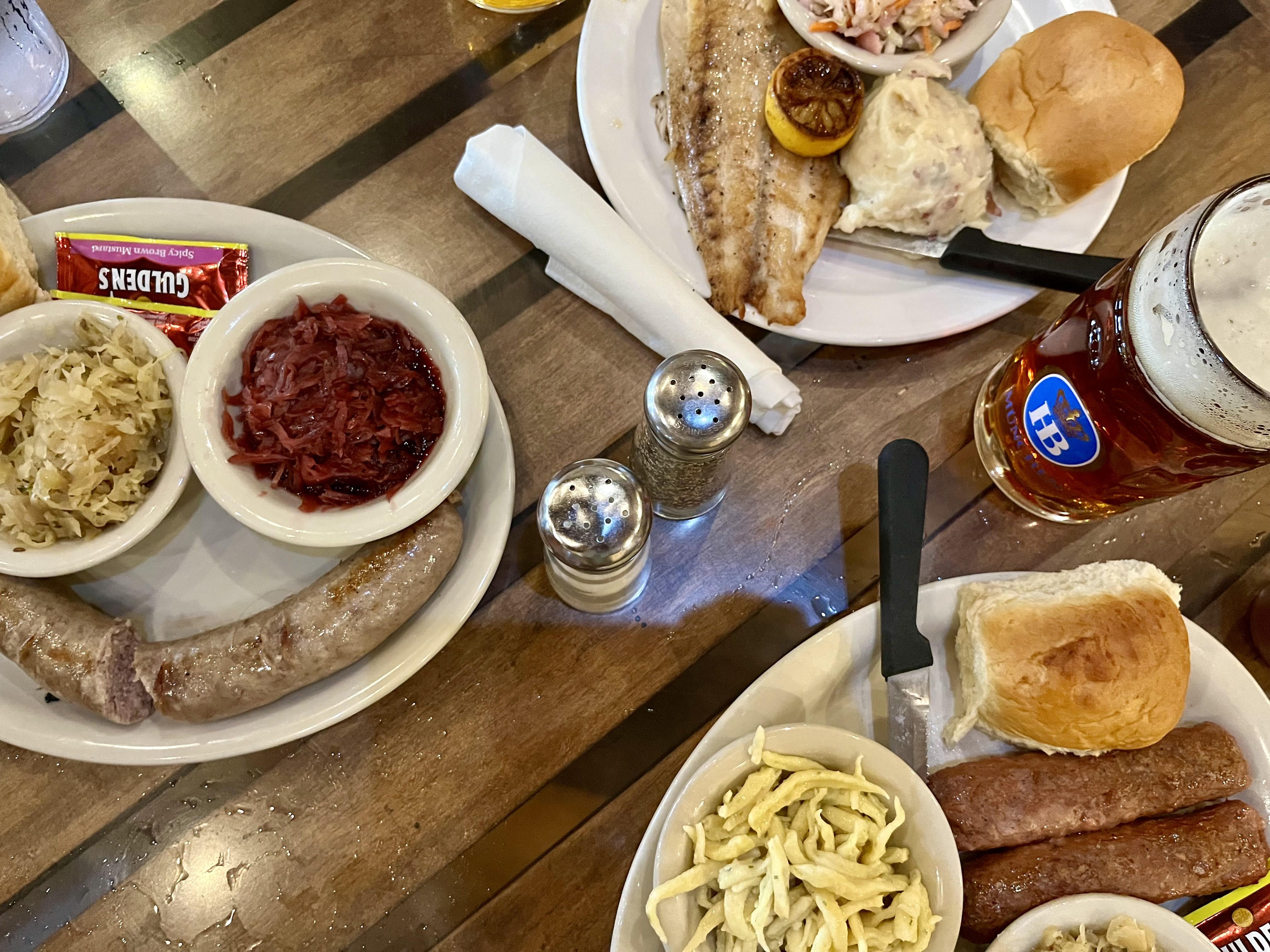 A wooden table displays German food like brats and grilled fish, with a large mug of beer.