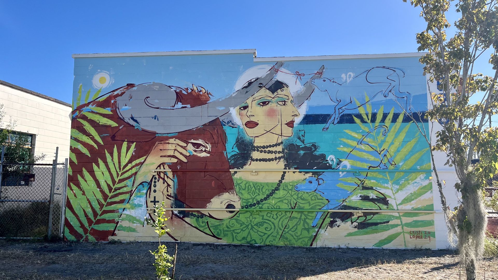 A mural of a woman with black hair and black beads looped around her neck looking at the viewer with her hand caressing a brown bull. The mural is decorated with palm fronds.