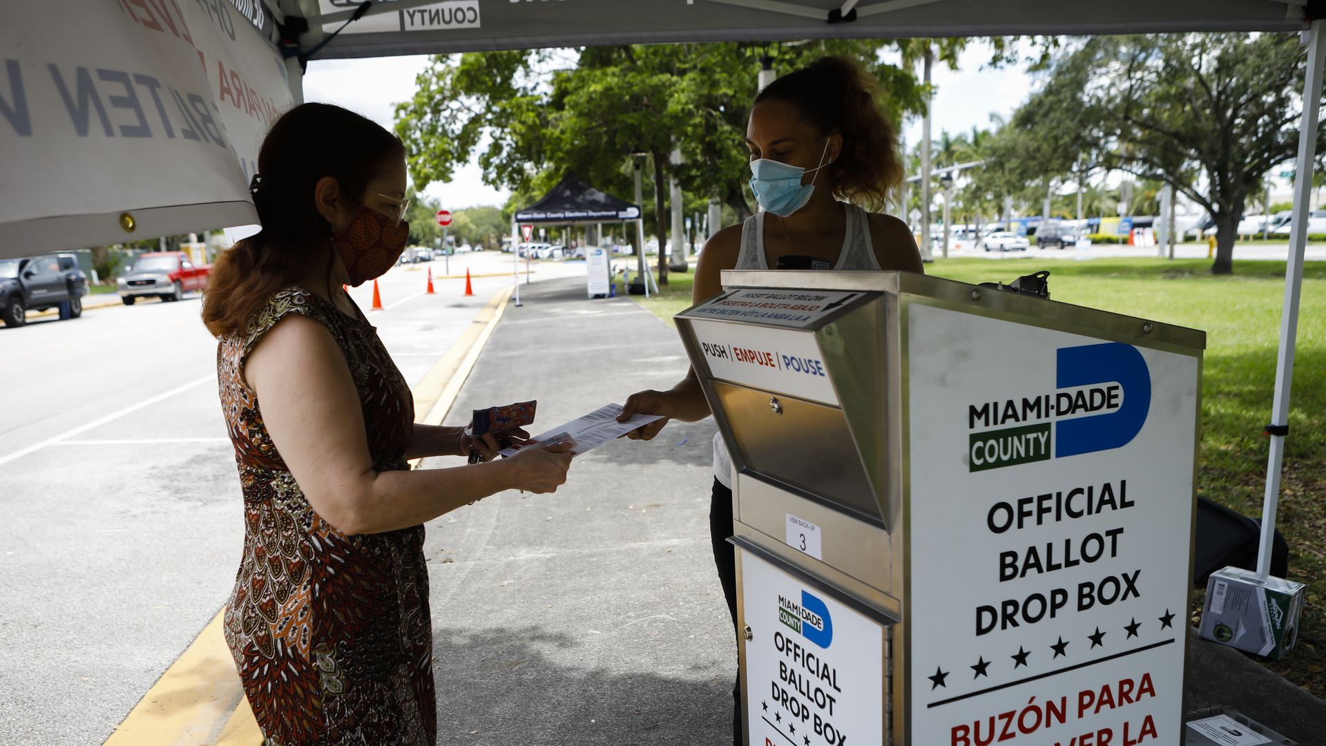 Photo of a person handing in their ballot to a voting booth worker next to a dropbox