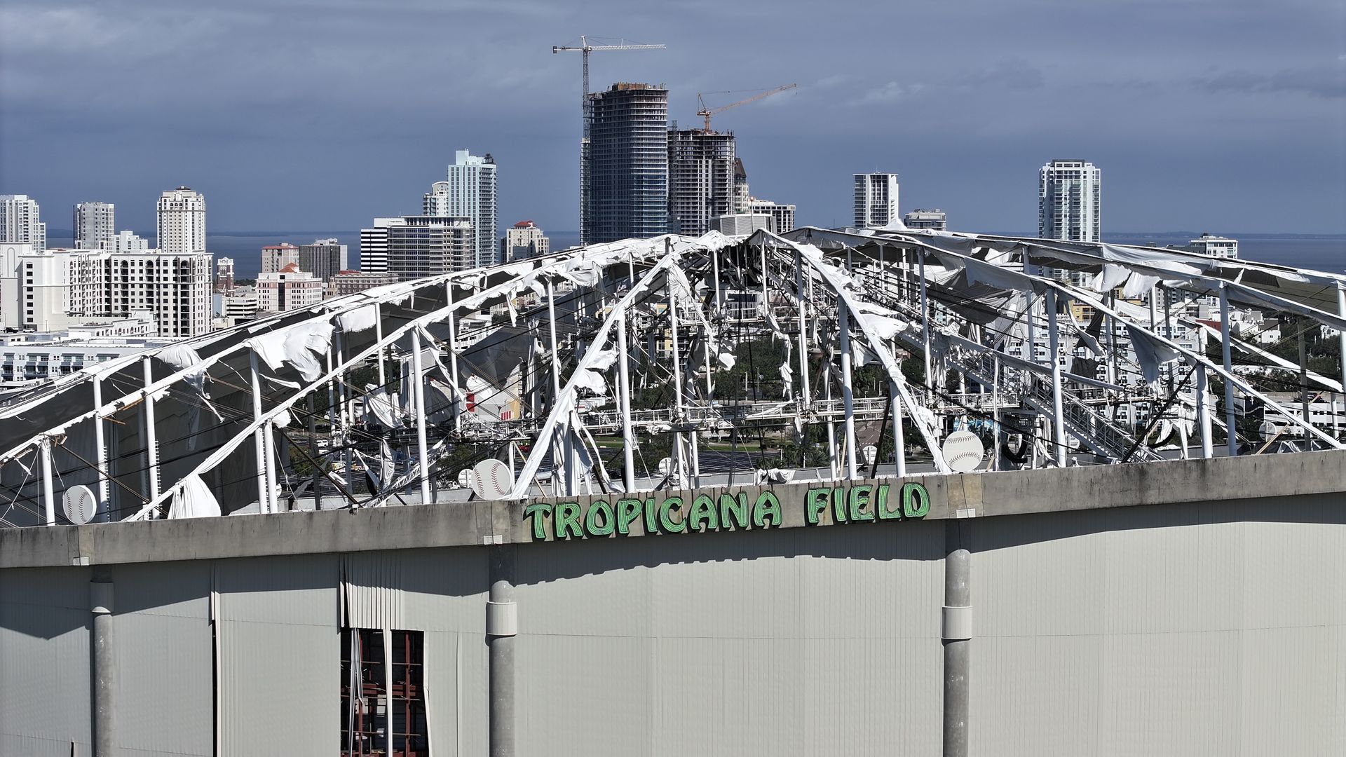 A dome with the roof ripped off with a sign that says "TROPICANA FIELD" in green letters. In the background, skyscrapers peak out from a skyline.