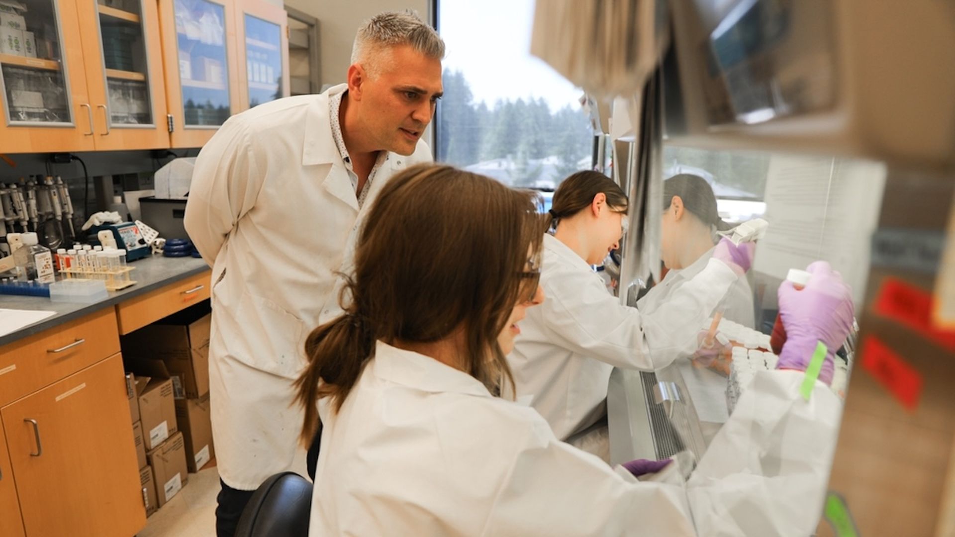 A photo of a person standing in a white lab coat, crouched over to view another person inserting a testing tube into something off screen.