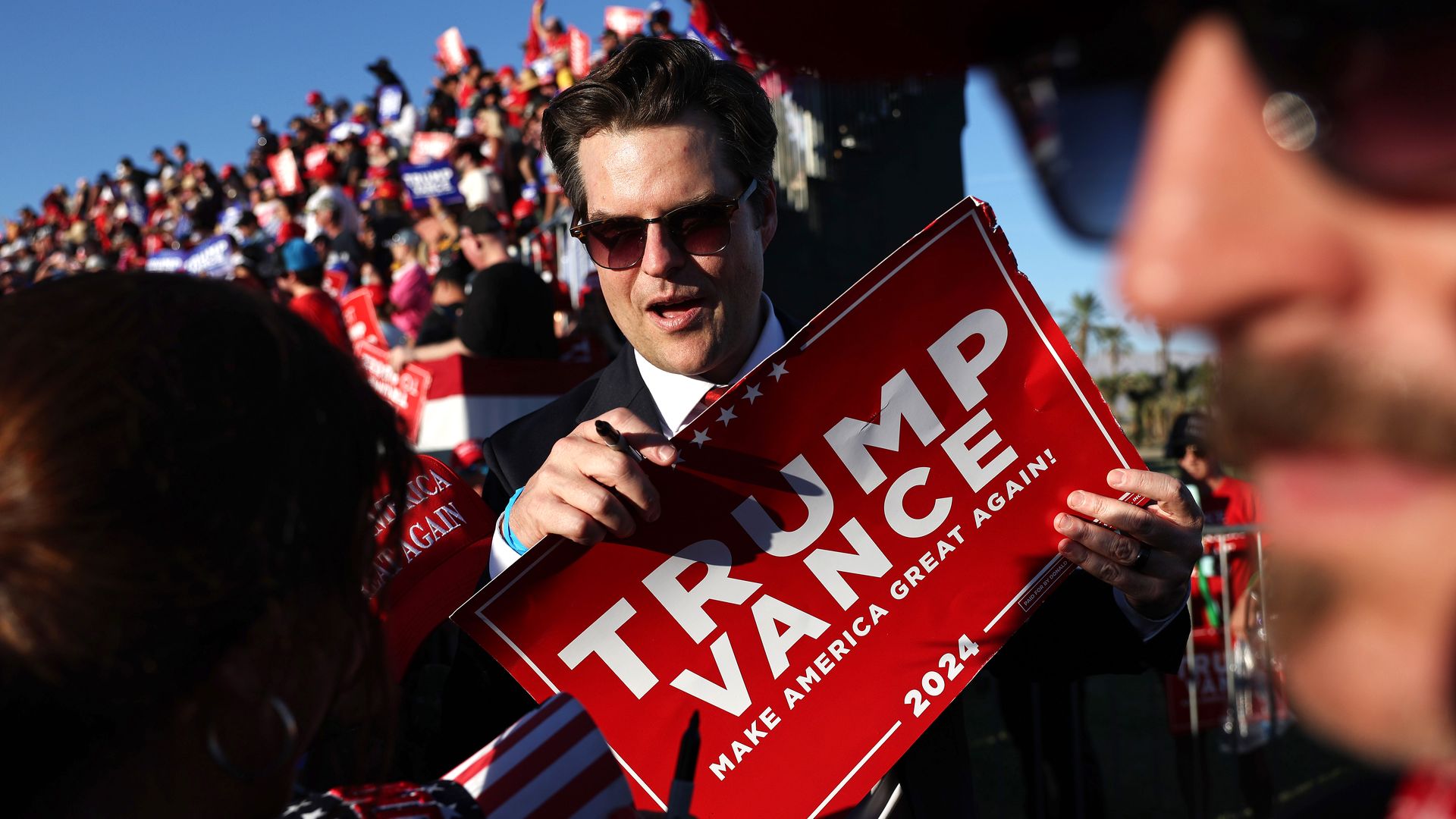 Rep. Matt Gaetz, wearing sunglasses and holding a red Trump Vance sign while flanked by a large crowd.