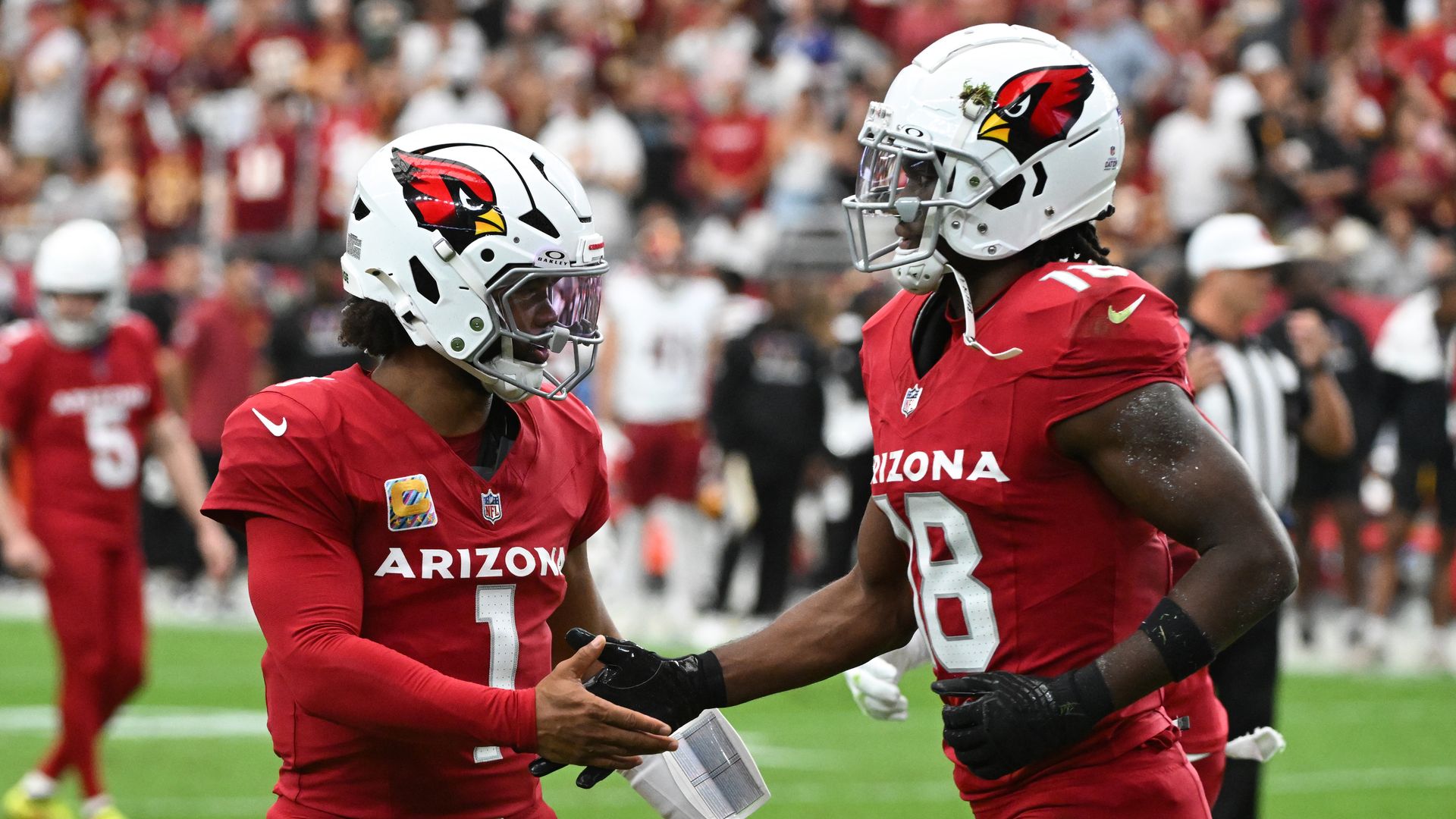 Kyler Murray and Marvin Harrison Jr., both wearing red Arizona Cardinals football uniforms with white helmets, shake hands on the field. 