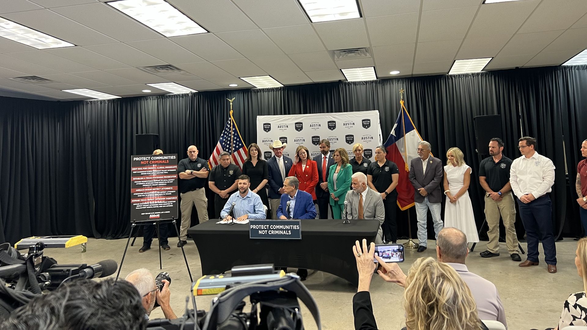 Wide view of a press conference: a row of officials on stage behind a banner that reads \"PROTECT COMMUNITIES NOT CRIMINALS\"; American and Texas flags flank the podium, photographers in the foreground.