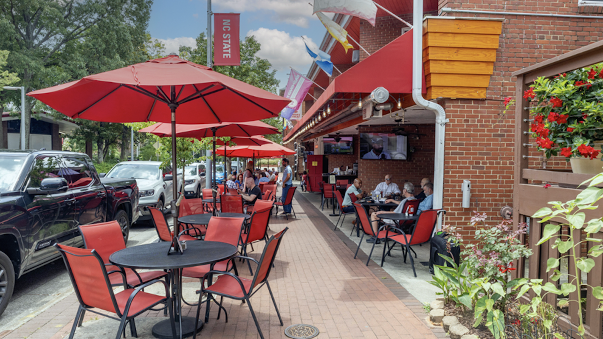Outdoor seating area at a brick-walled restaurant with red umbrellas and chairs, customers dining, NC State banner above, parked cars along the street, and flowers in a small garden.