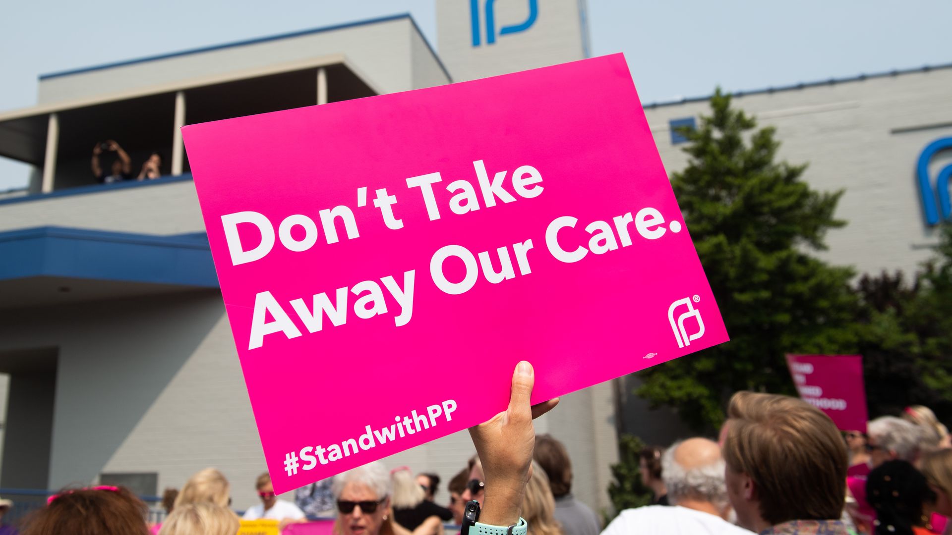 Picture of a sign that says "don't take away our care" in front of a Planned Parenthood clinic