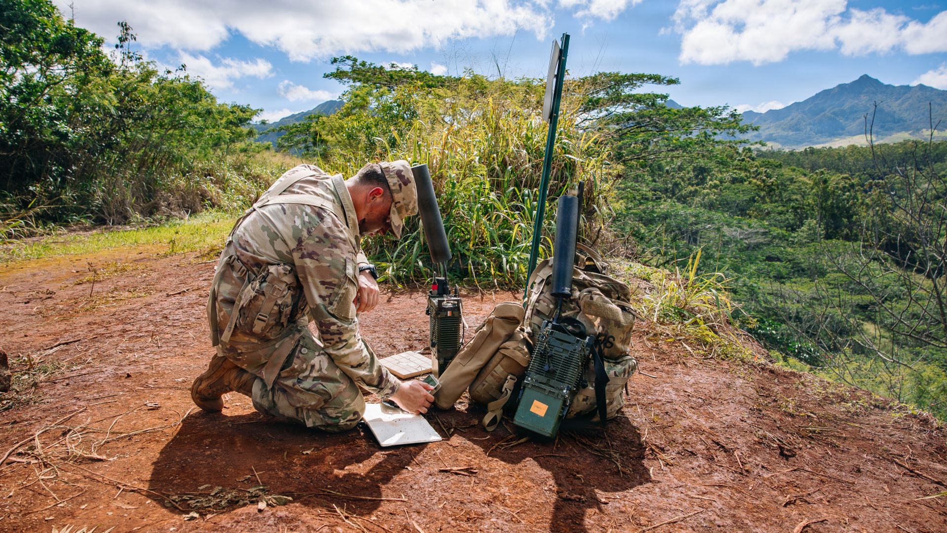 Soldier in camouflage uniform kneels on dirt ground near radio equipment and backpack amid green trees and mountains under a partly cloudy blue sky.