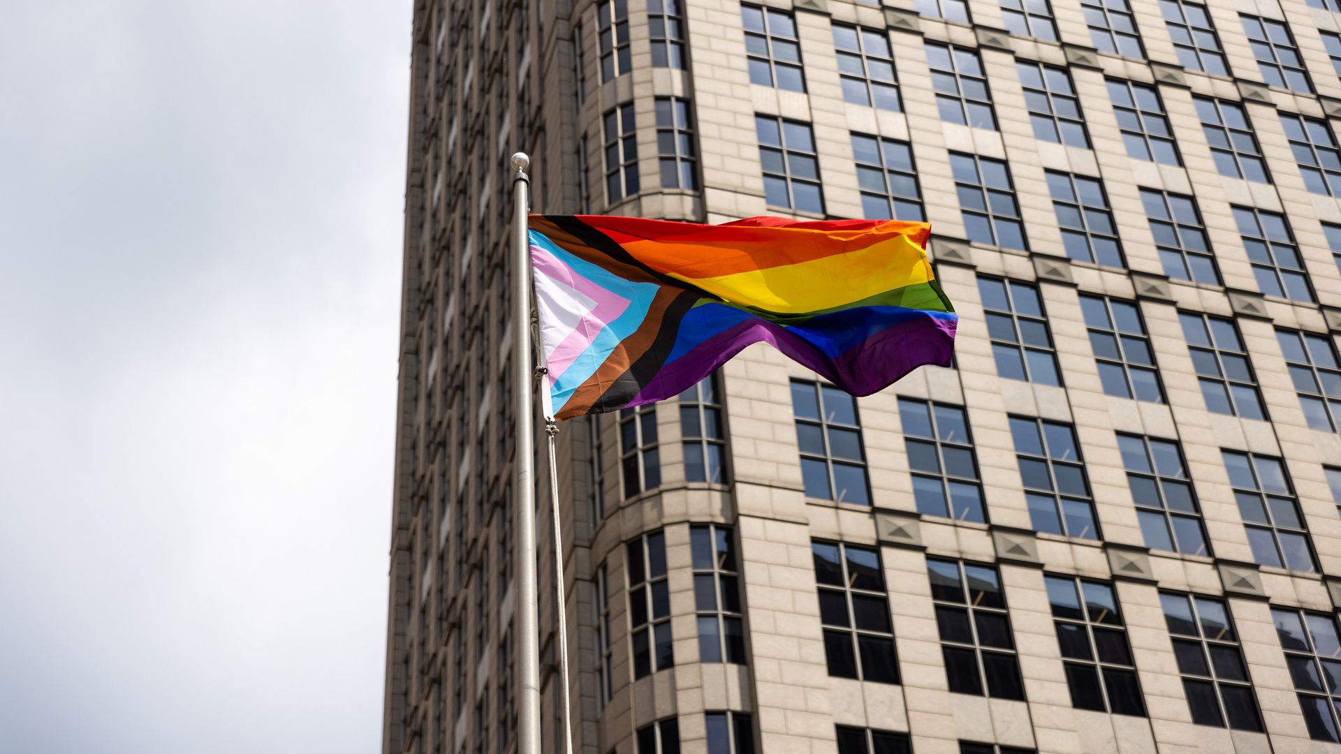 A Pride flag blows in the wind in Detroit in front of a high-rise building.