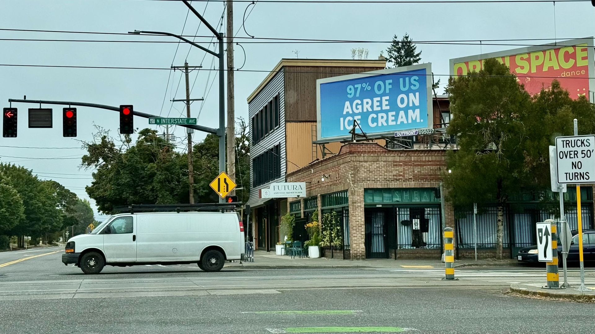 Urban intersection with a white cargo van, traffic lights, and overhead power lines. A blue billboard reads "97% OF US AGREE ON ICE CREAM." A brick building, trees, and a pink sign in the background.