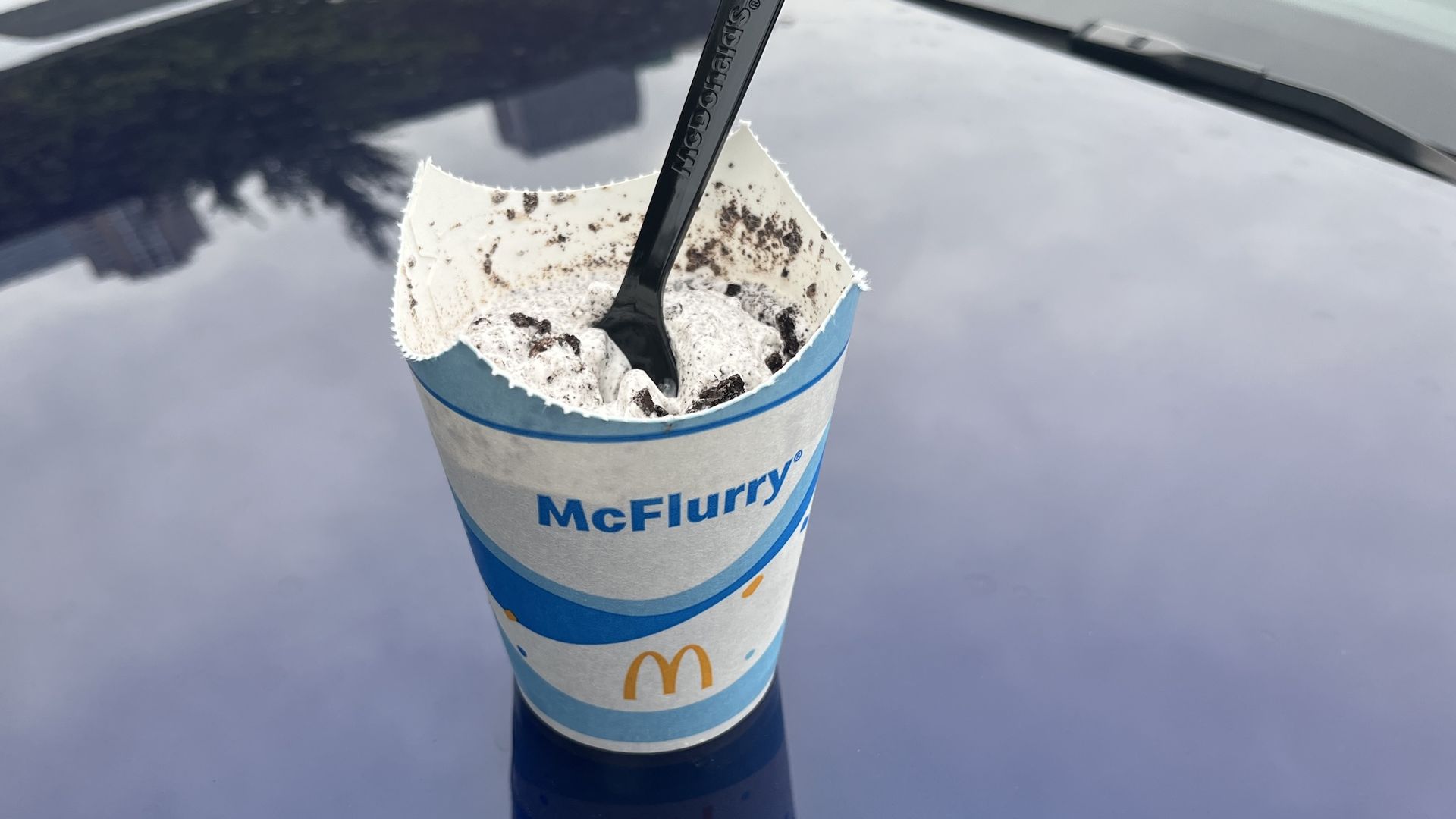 Blue and white paper cup that reads McFlurry with ice cream with Oreos with a black plastic spoon.