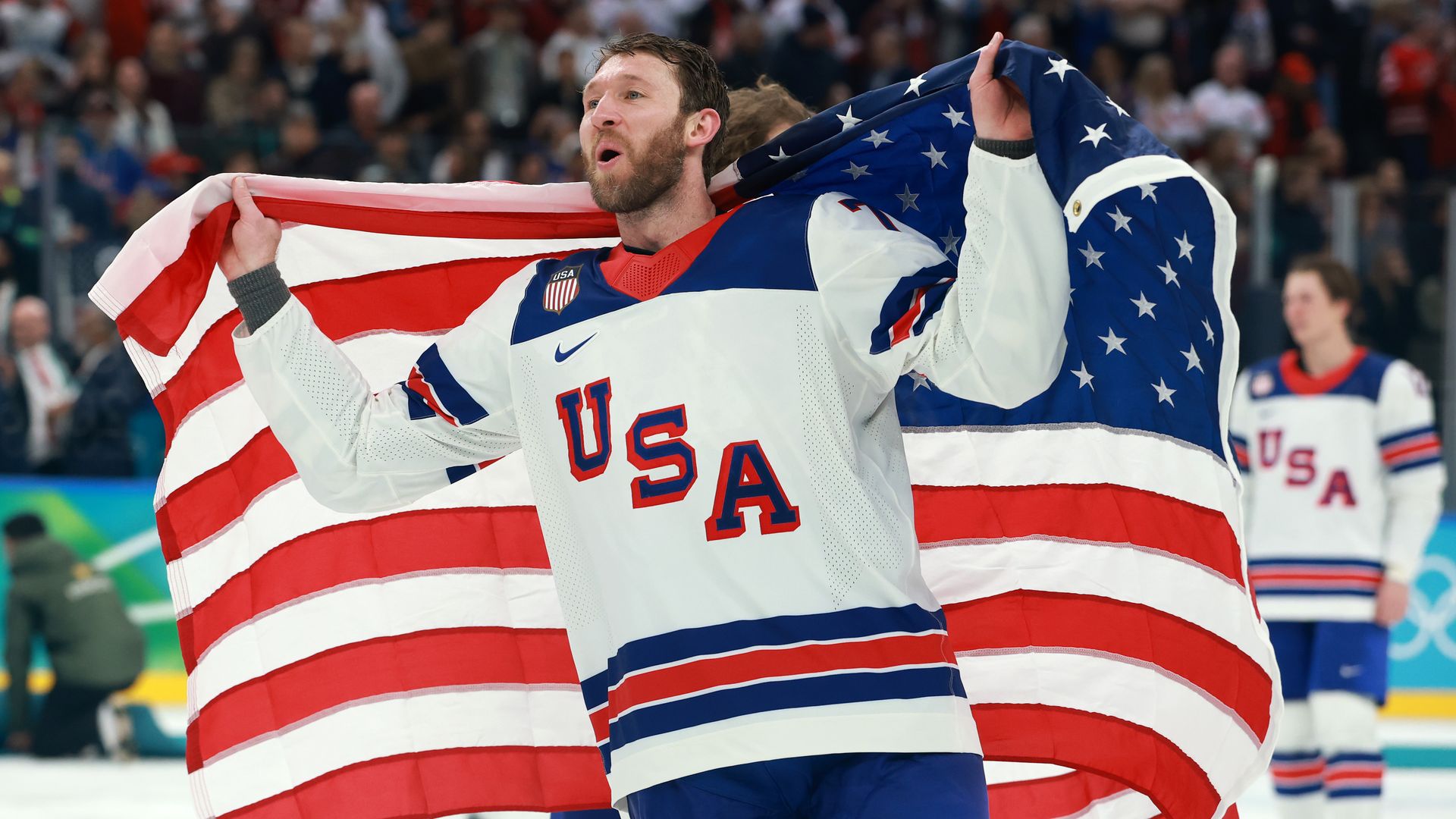 Hockey player in white USA jersey holding American flag behind him on ice rink with crowd in background, celebrating.