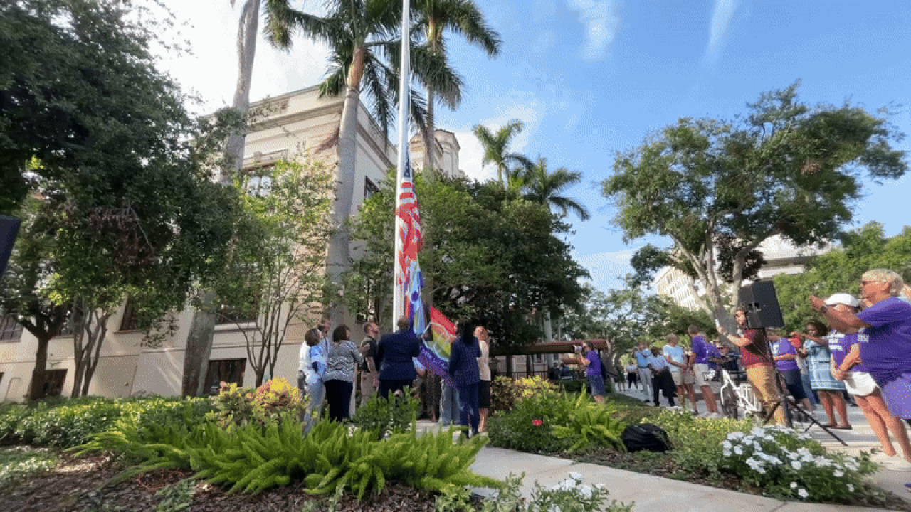 A group of people raise an American flag, city of St. Petersburg, FL, flag and rainbow St. Pete Pride flag in front of a historic building.