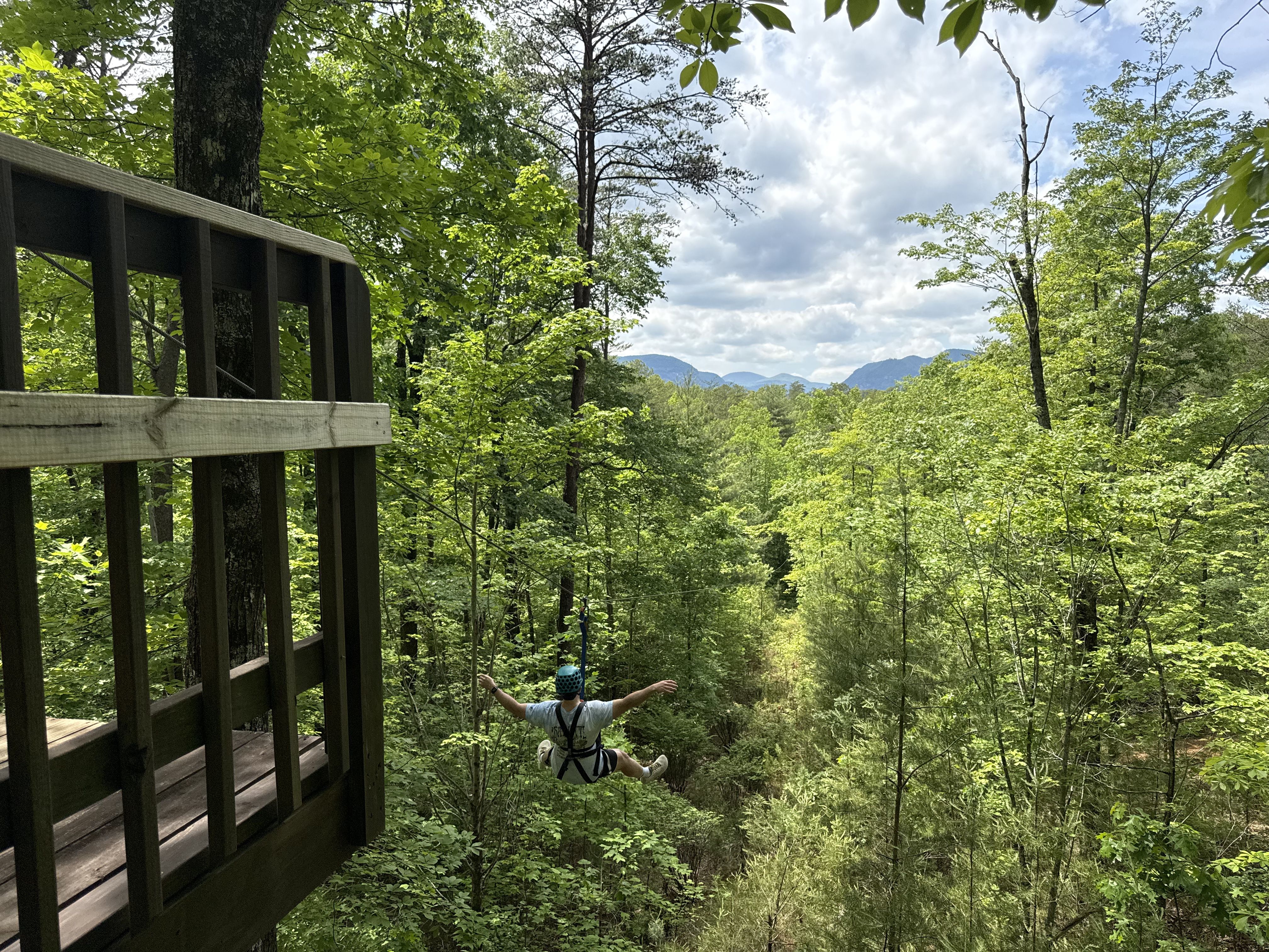 Forest setting with a wooden railing deck on the left, lush green trees all around, and distant blue mountains under a partly cloudy sky. A person in a helmet and harness zip-lines through the woods.