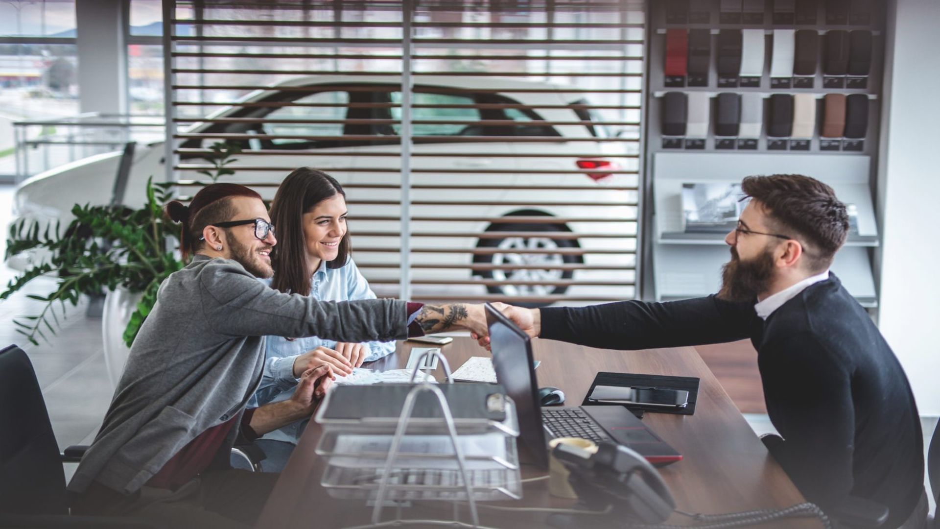 Man and woman sitting at a table shaking hands with a bearded man across from them in a modern office with a white car and fabric samples behind glass panels.