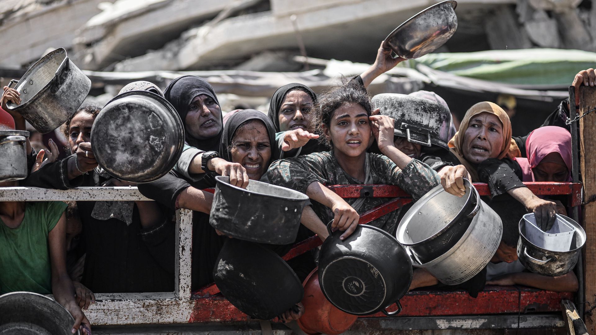 A group of women in headscarves urgently hold up pots and pans, reaching out from behind a rusted metal barrier in a crowded, dusty outdoor setting.
