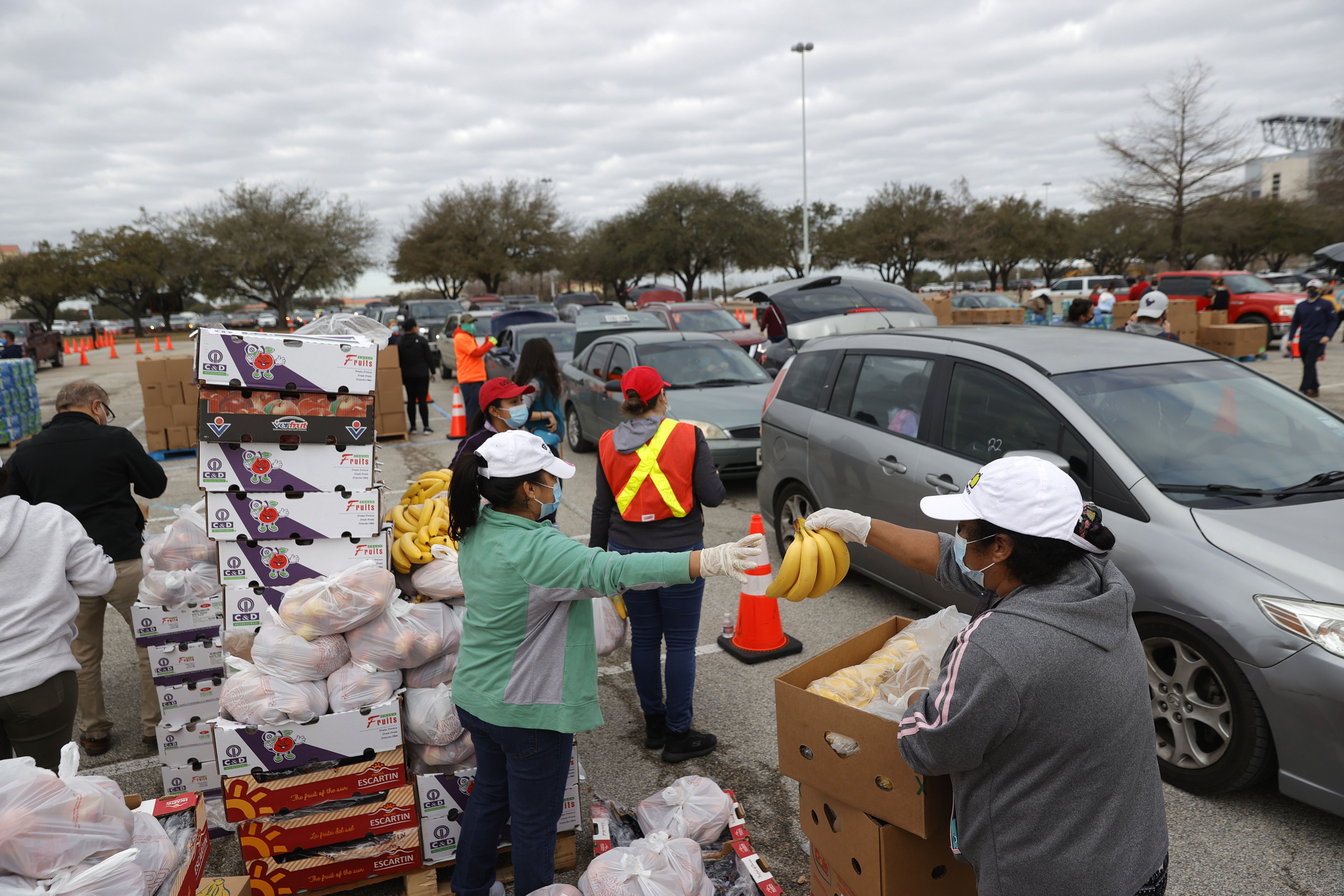 A line of cars in a parking lot, as volunteers wearing bright vests hand out boxes of food