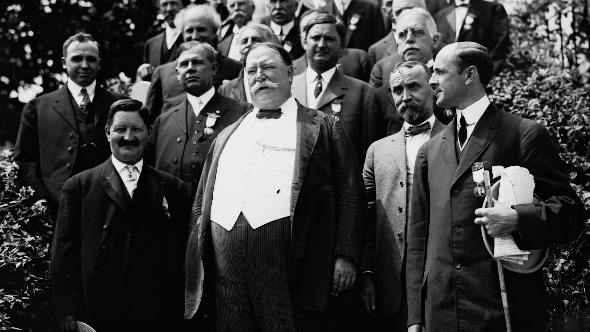 Black and white photo of President Taft and a large group of men wearing early 20th-century formal attire, including suits, bow ties, and medals, posing outdoors in front of greenery and a building.