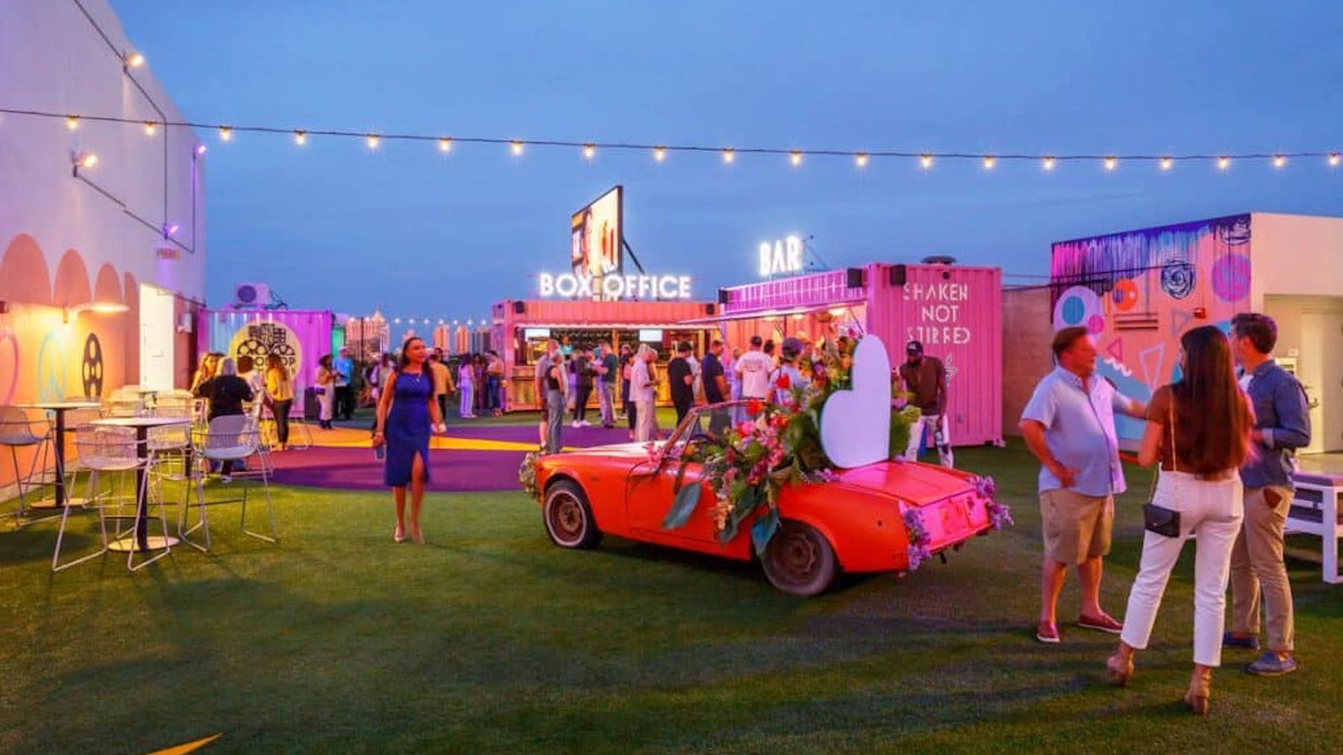 At dusk, twinkle lights hang from a rooftop while people mingle on artificial grass