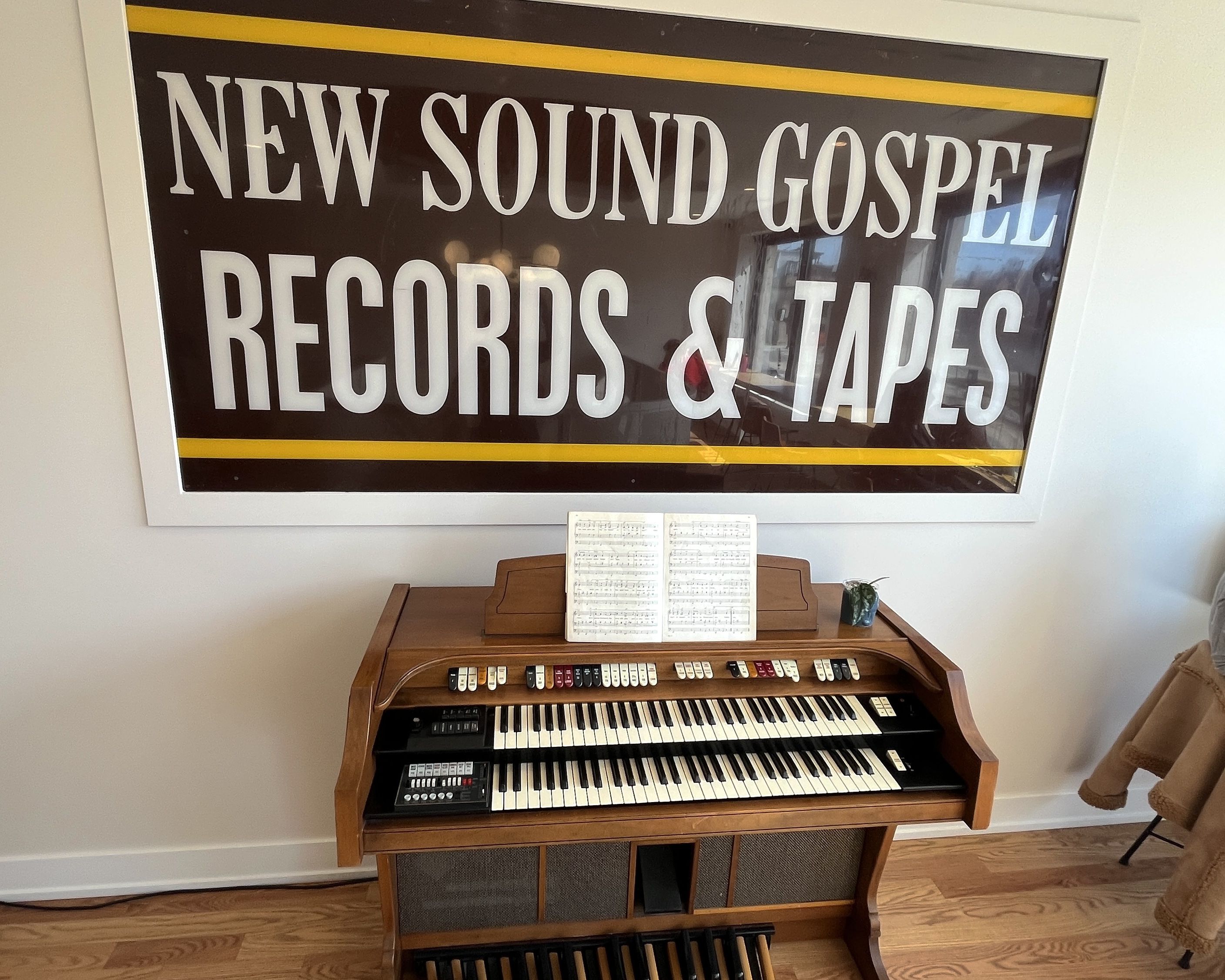 Large framed sign reads 'NEW SOUND GOSPEL RECORDS & TAPES' in white on black with yellow borders; below is a wooden organ with two keyboards, sheet music, and a small potted plant.