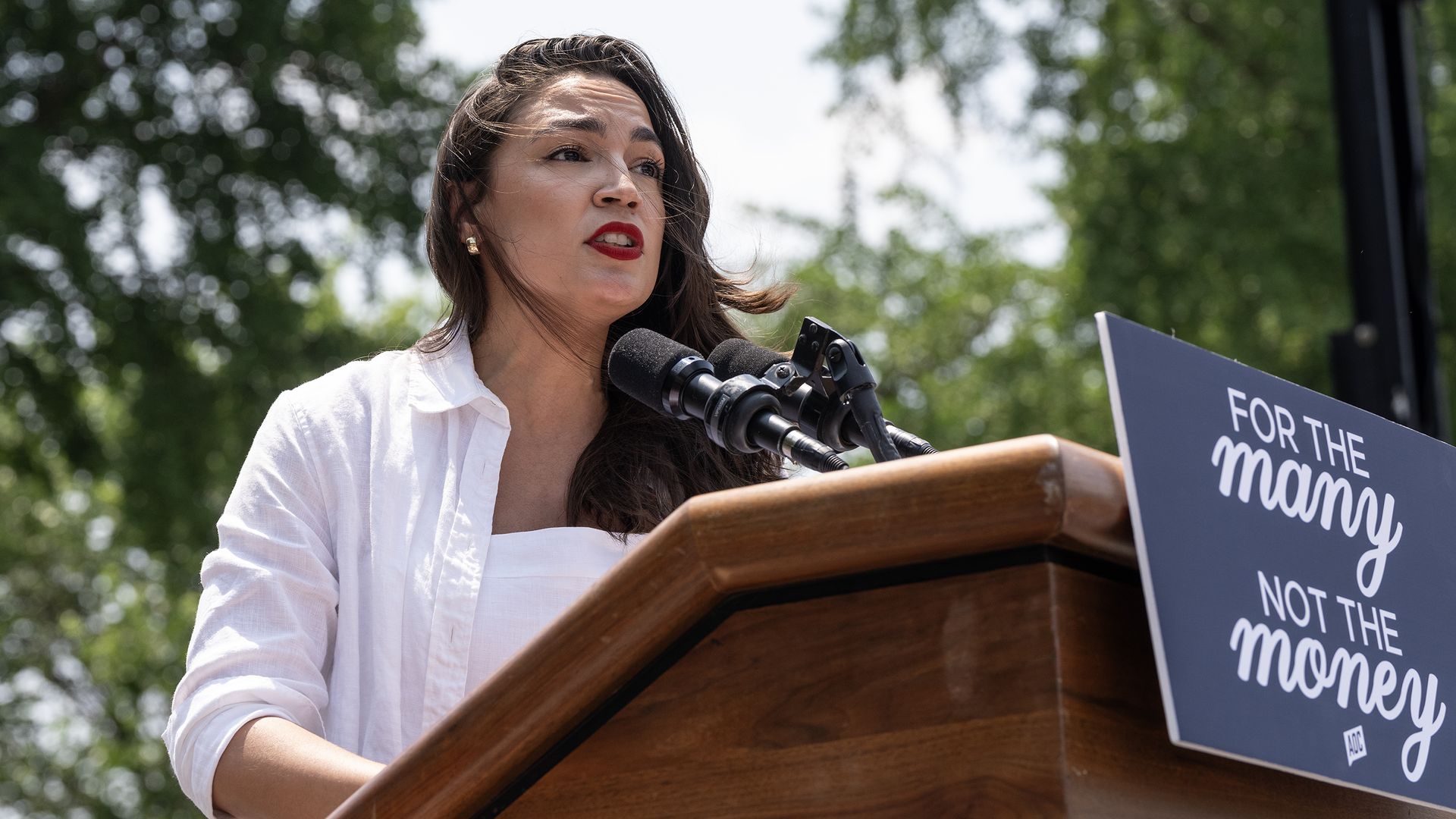 Rep. Alexandria Ocasio-Cortez wearing, a white blouse and speaking at a wooden podium in front of trees.
