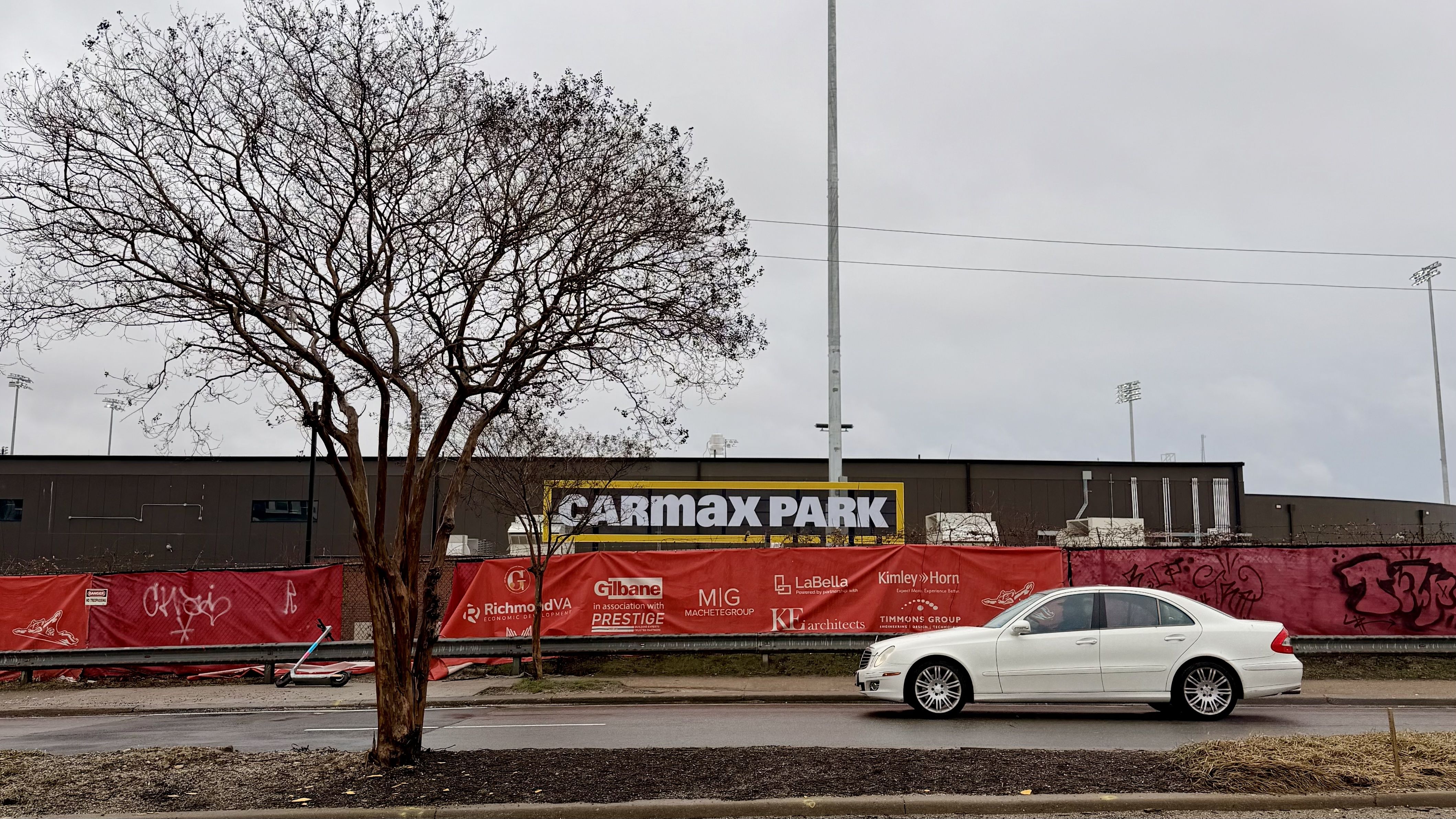 White sedan parked on street in front of bare tree, red construction banners with logos, and large brown building with "Carmax Park" sign under overcast sky.