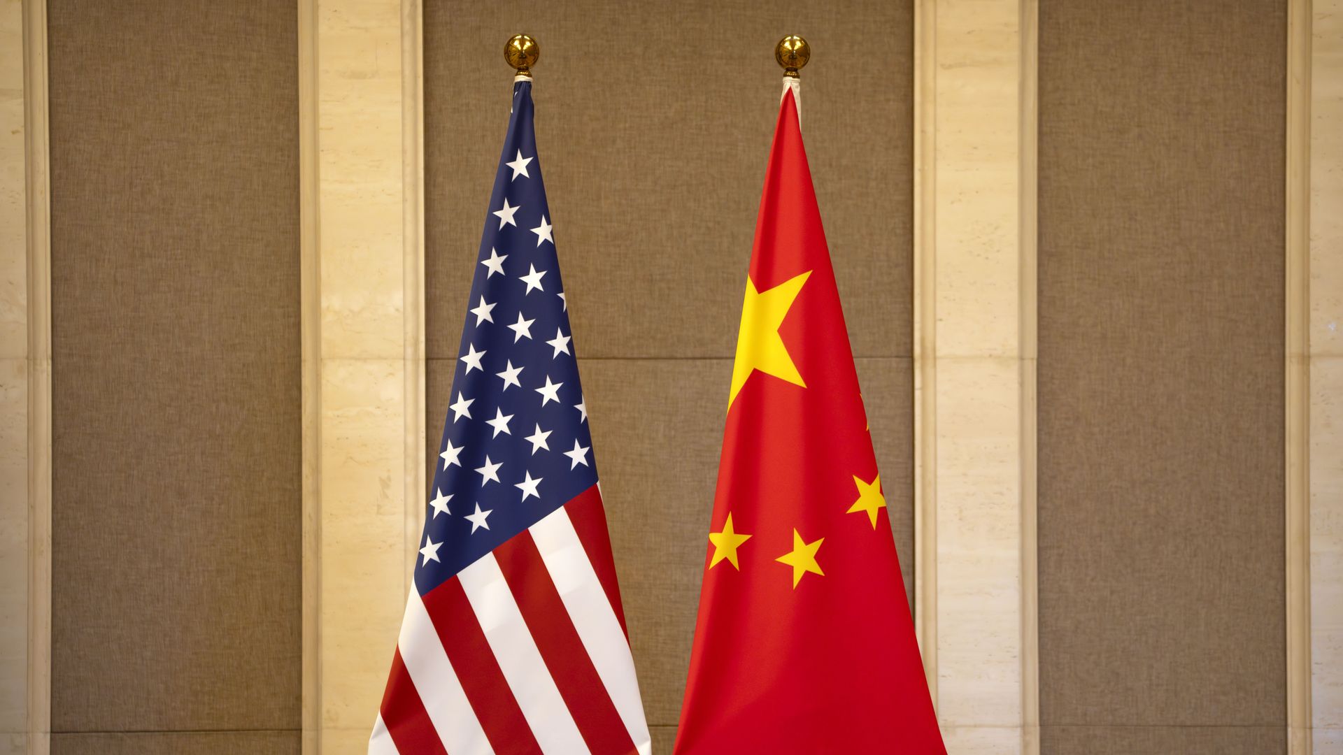 Side-by-side flags of the United States with stars and stripes, and China with a large yellow star and four smaller stars on a red background, against beige and brown wall panels.