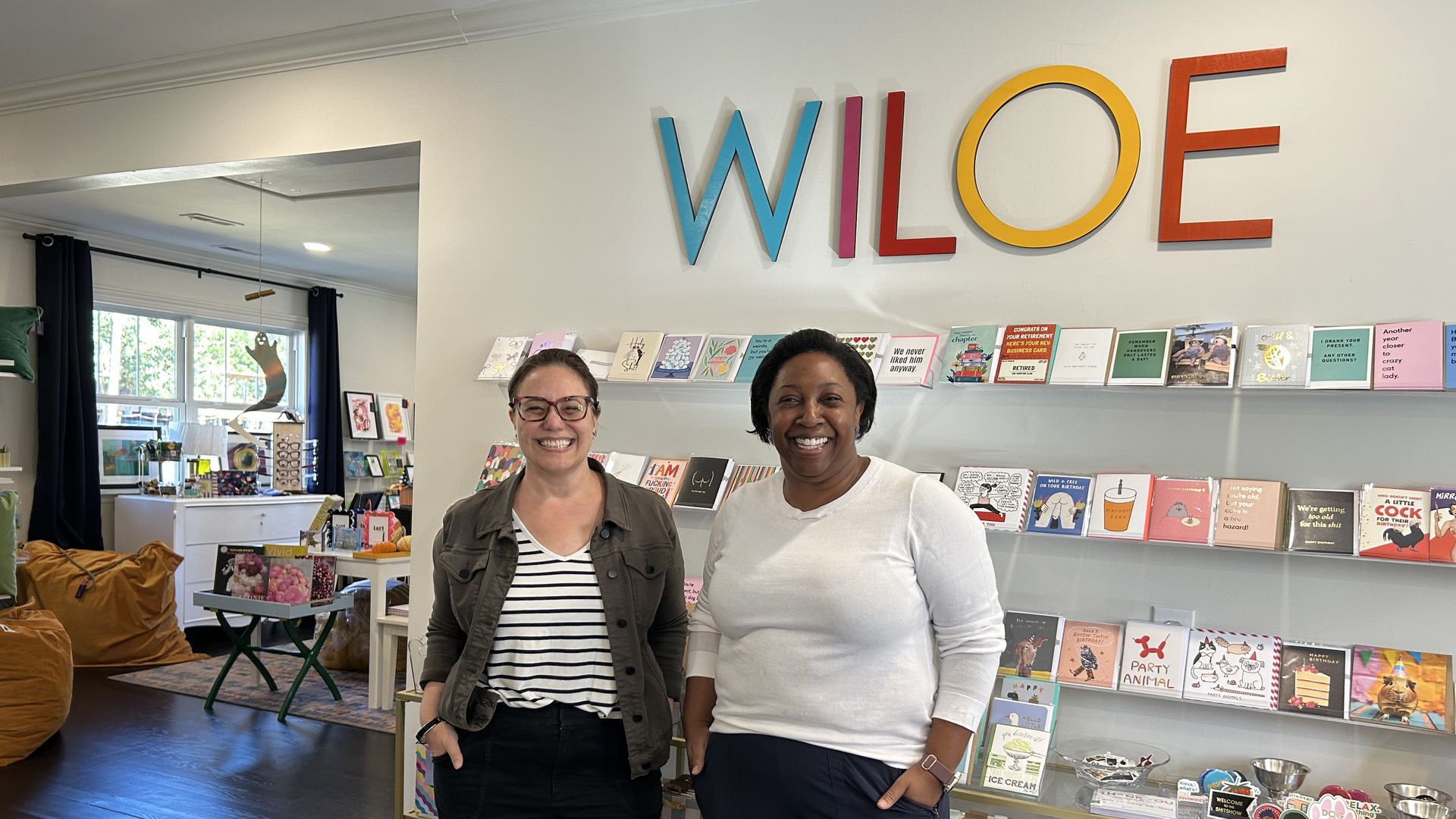 Two smiling women stand in a colorful store with greeting cards and gifts under a large sign reading WILOE in blue, red, yellow, and orange letters on the wall behind them.