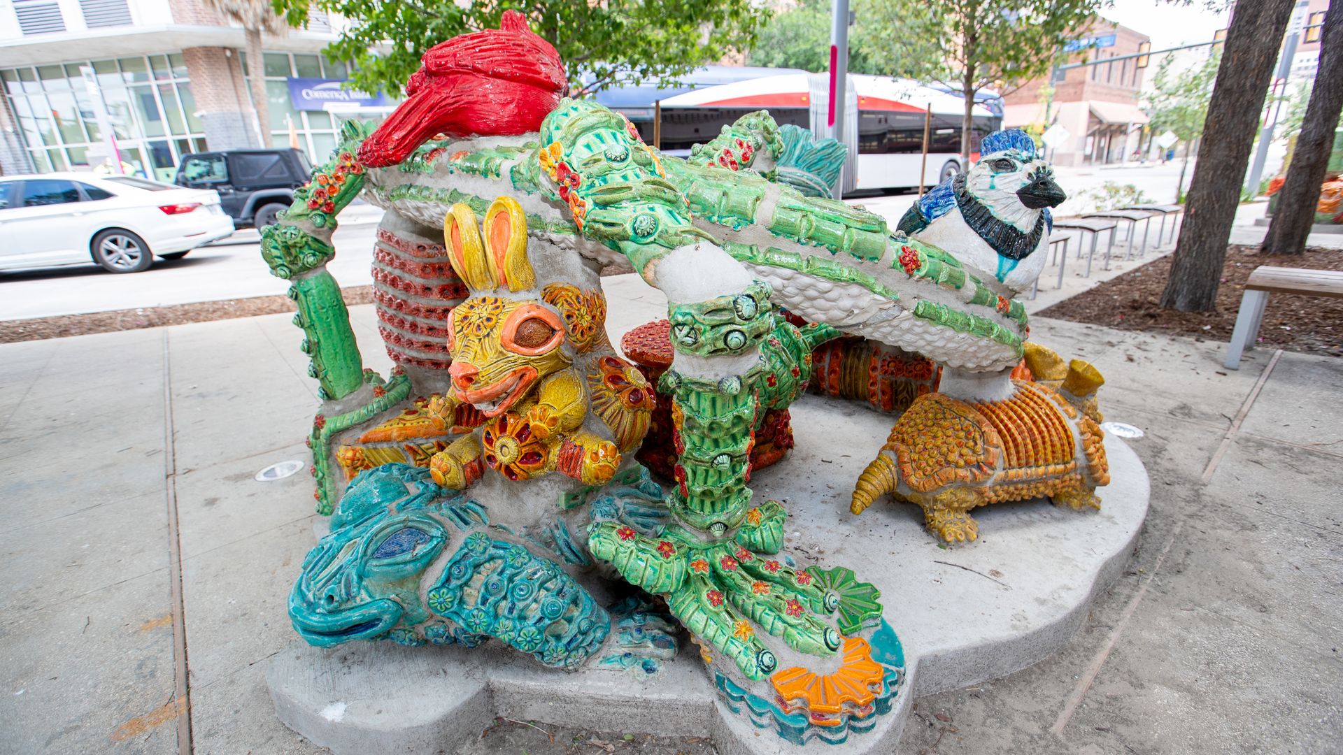 A colorful collection of clay animal sculptures includes a yellow rabbit, a blue bird and more alongside a downtown San Antonio street with a bus in the background. It is part of “Creatures of the Creek: A Fragile Balance” by Diana Kersey.