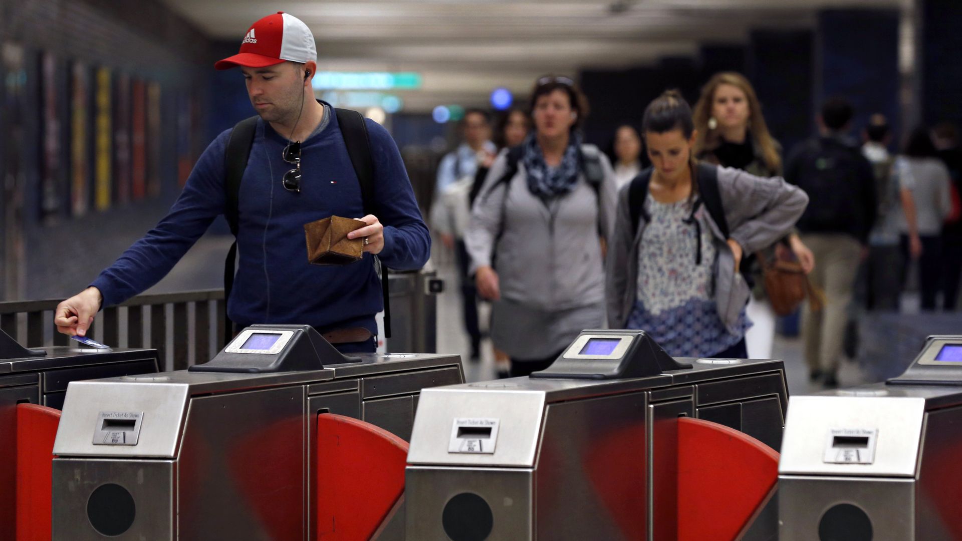 Man in red and white cap uses a ticket to pass subway turnstile with red barriers, while people walk nearby in a busy underground station.