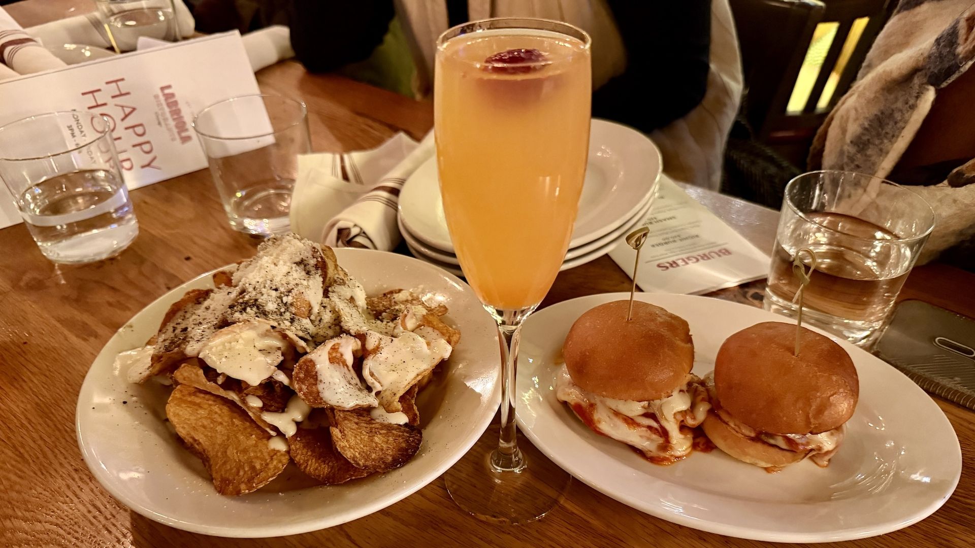Table with a plate of crispy chips topped with white sauce and seasoning, two small chicken sliders on a white plate, and a tall orange cocktail garnished with a cherry.