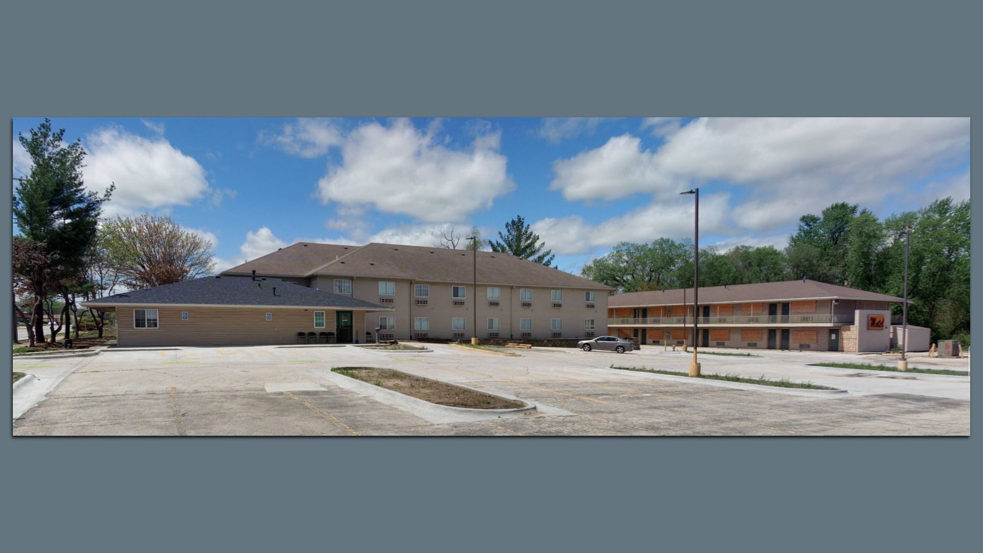 Wide view of a small motel with beige and gray buildings under a blue sky with white clouds, surrounded by trees and an empty concrete parking lot with a single car.
