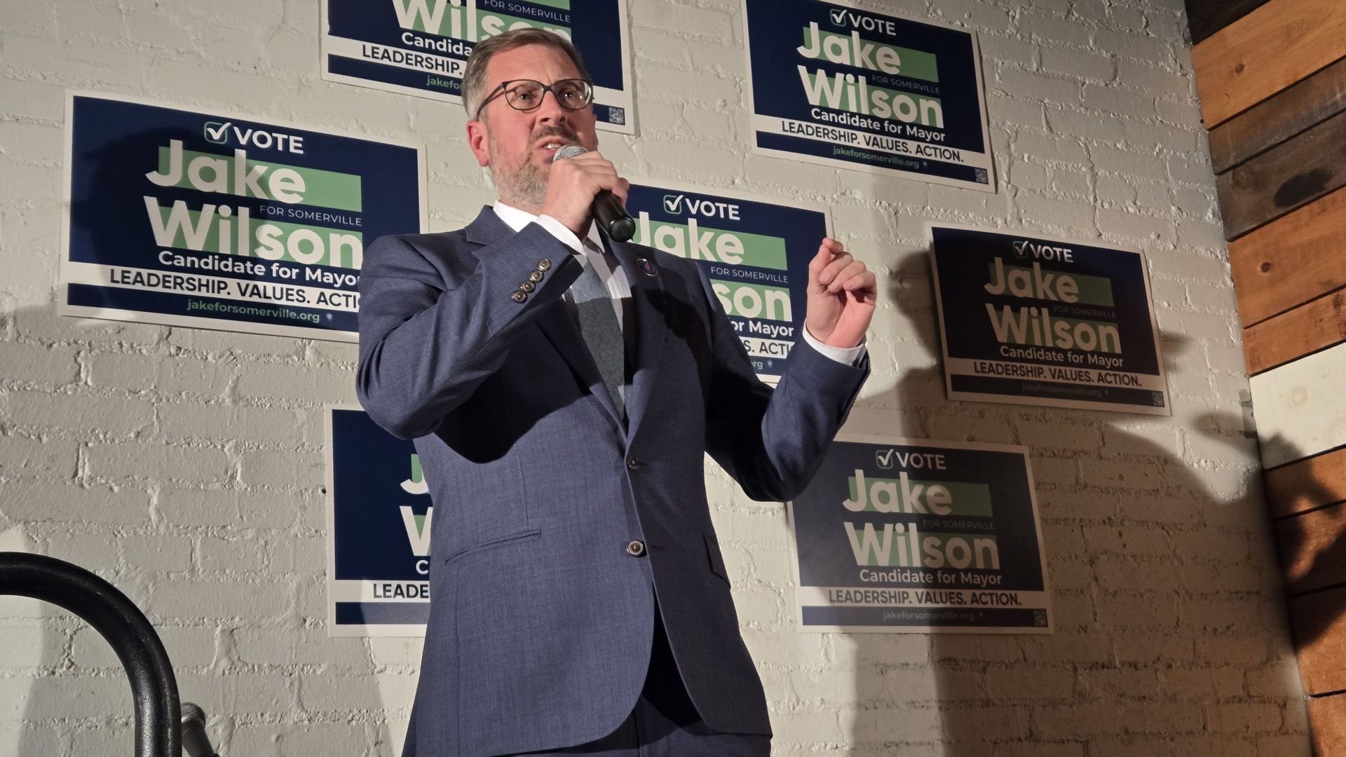 Man in blue suit and glasses speaking into a microphone in front of a white brick wall with multiple "Vote Jake Wilson for Mayor" campaign posters in blue and green colors.