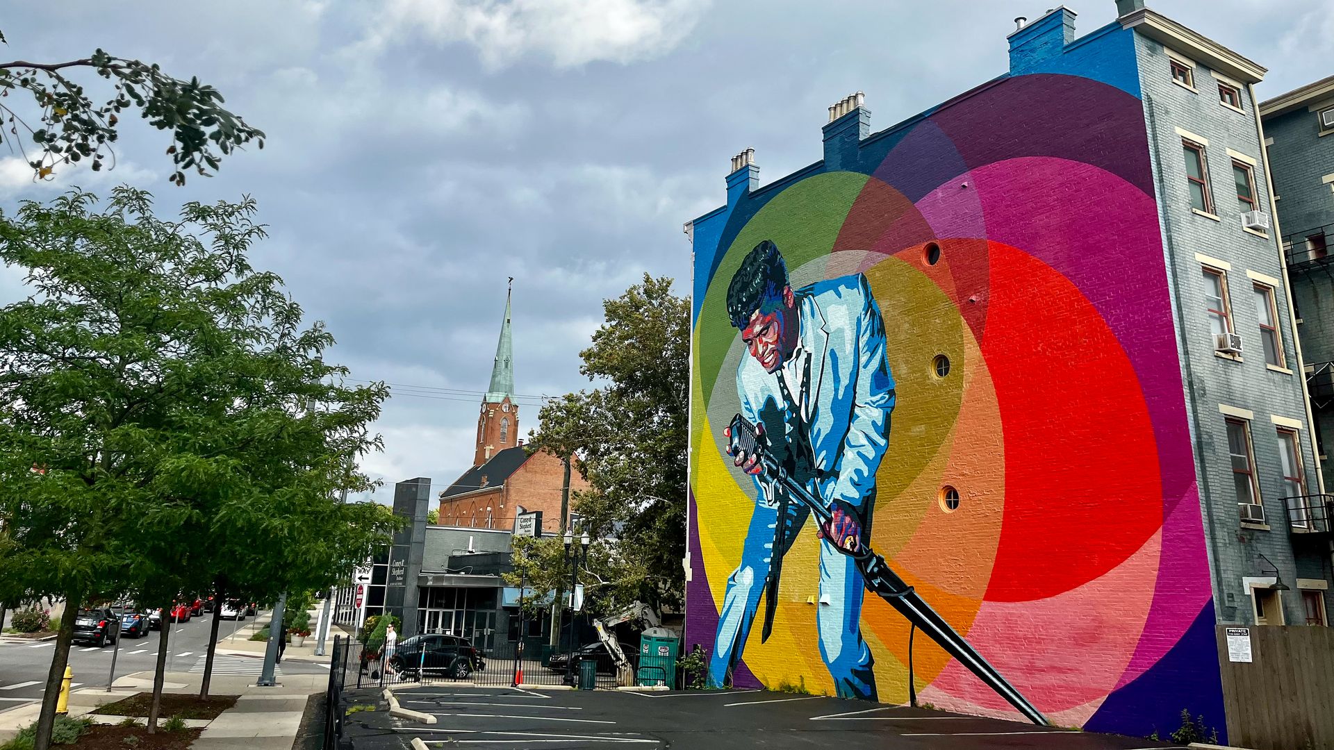 Urban street mural on a brick building featuring a stylized guitarist in blue tones, surrounded by overlapping rainbow arcs. Cloudy sky, trees, and a church steeple in the background.
