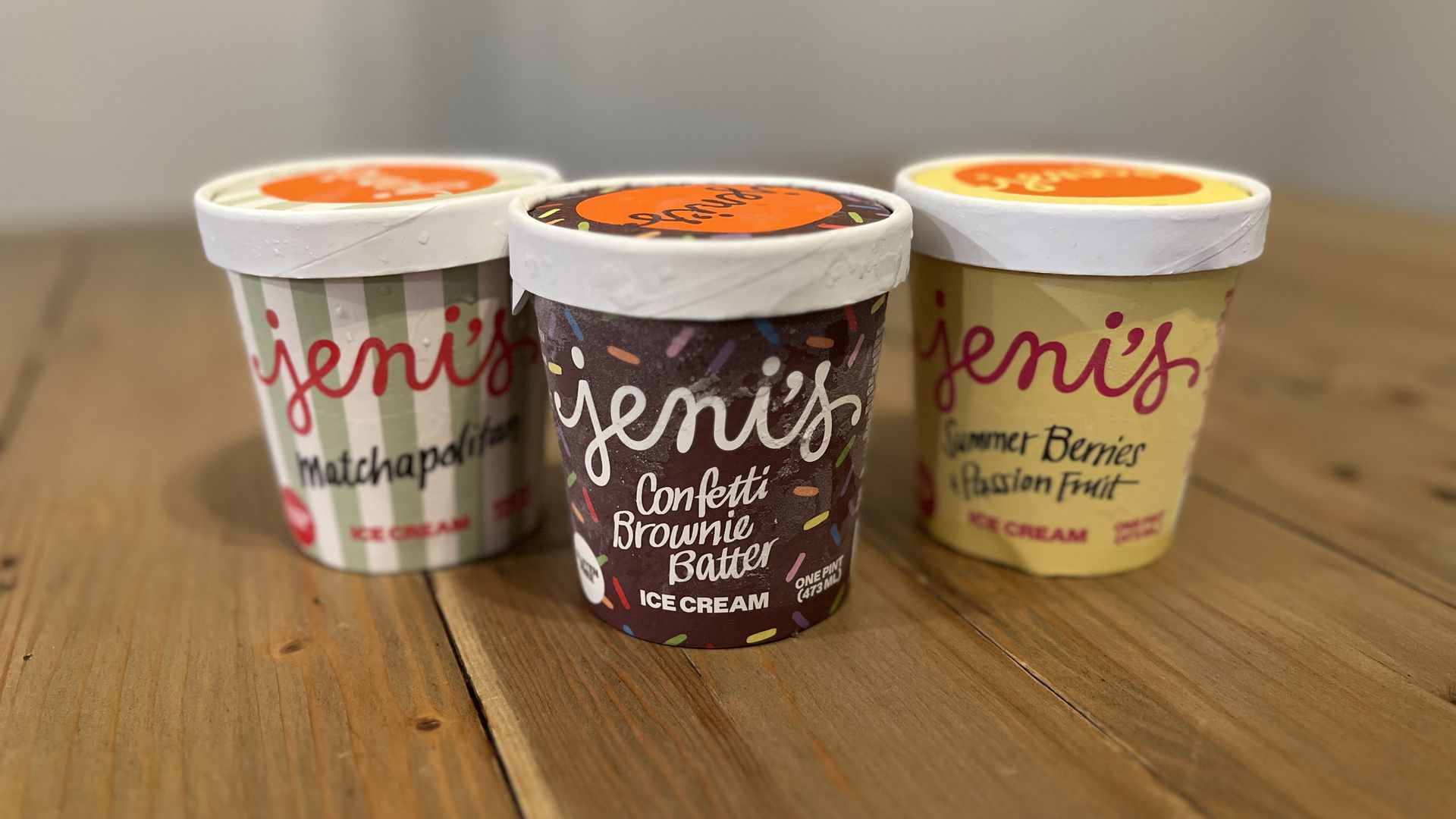 Three ice cream cups with colorful wrappers sit on a wooden table; center cup shows a purple confetti pattern, flanked by striped green and yellow cups, with a softly blurred background.