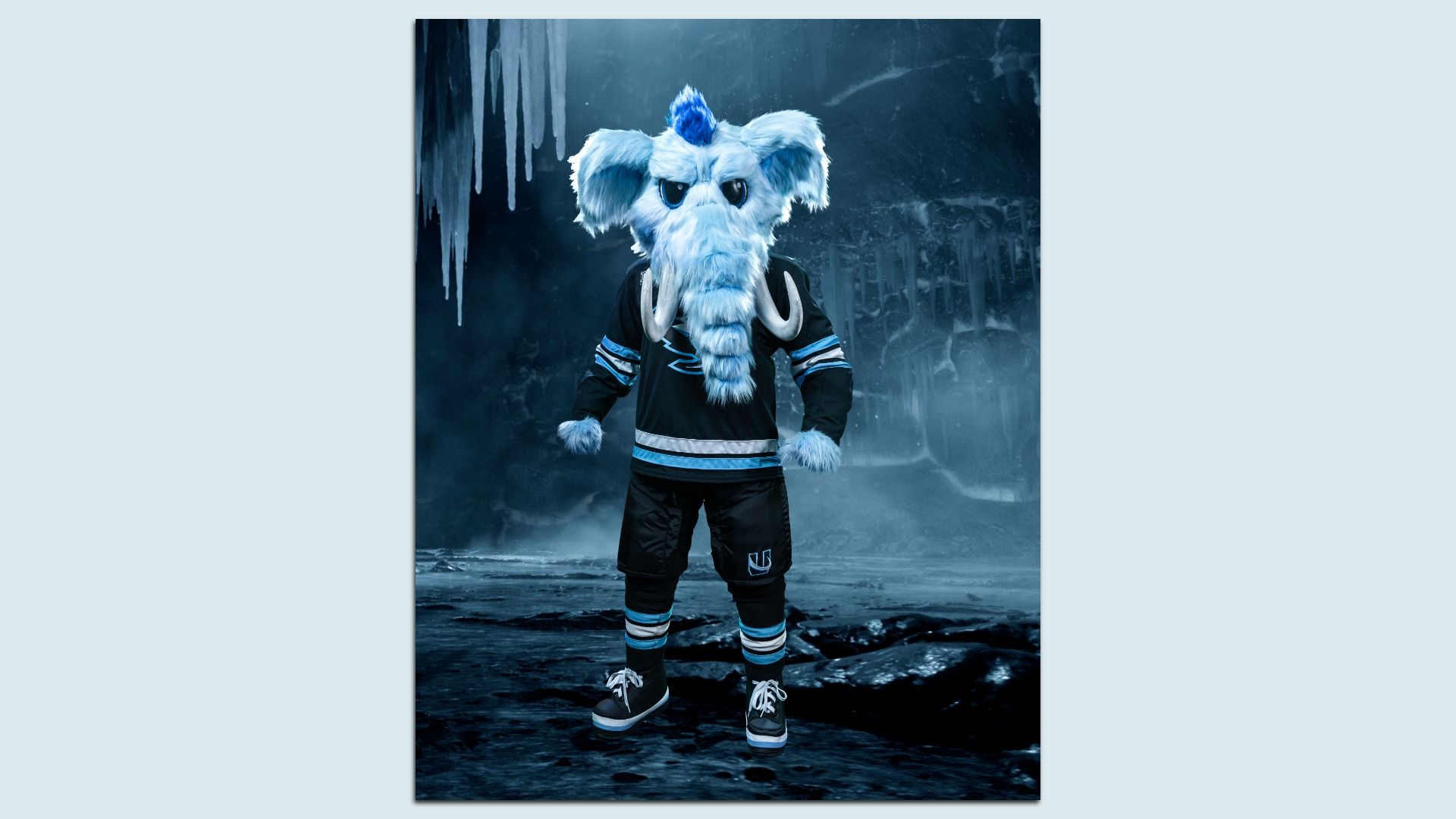 Blue and white furry mammoth mascot, named Tusky, with tusks, wearing a black and blue hockey jersey, standing in an icy cave with icicles hanging in background.