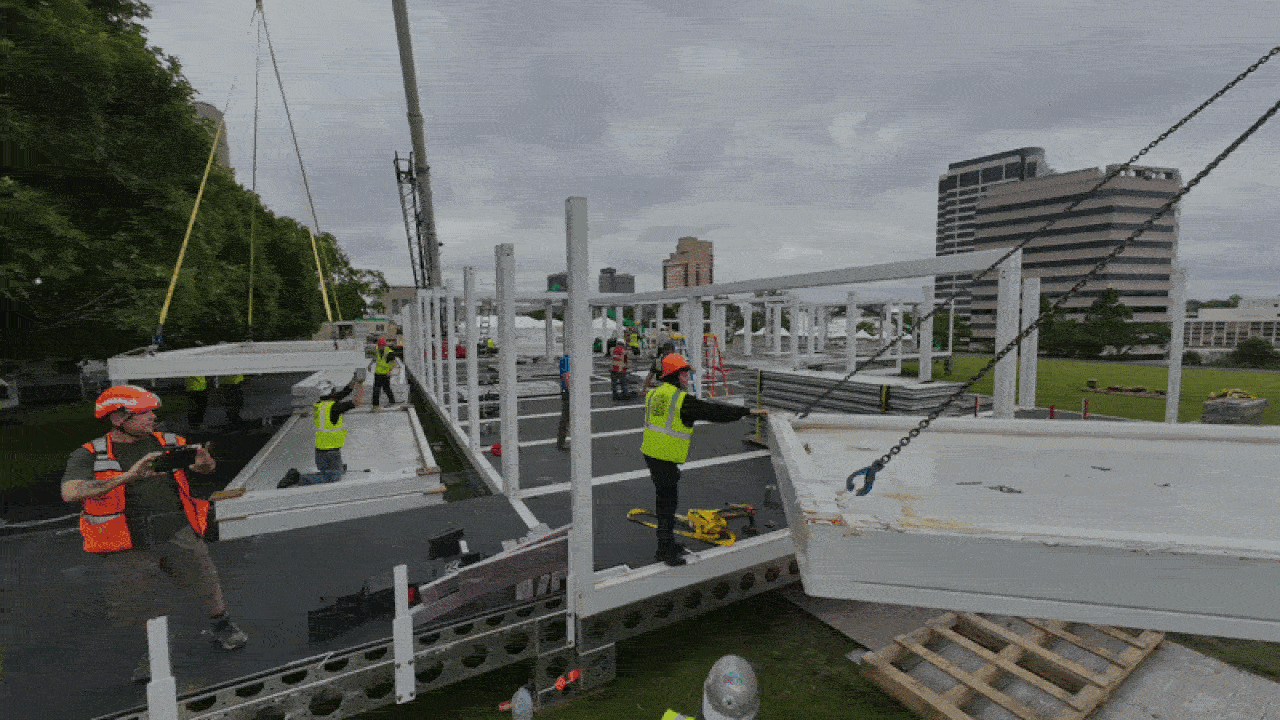 Construction site with a white steel frame; workers in orange helmets and neon vests position large white beams. Cranes, chains, and scaffolding surround the area, with a city skyline in the background.
