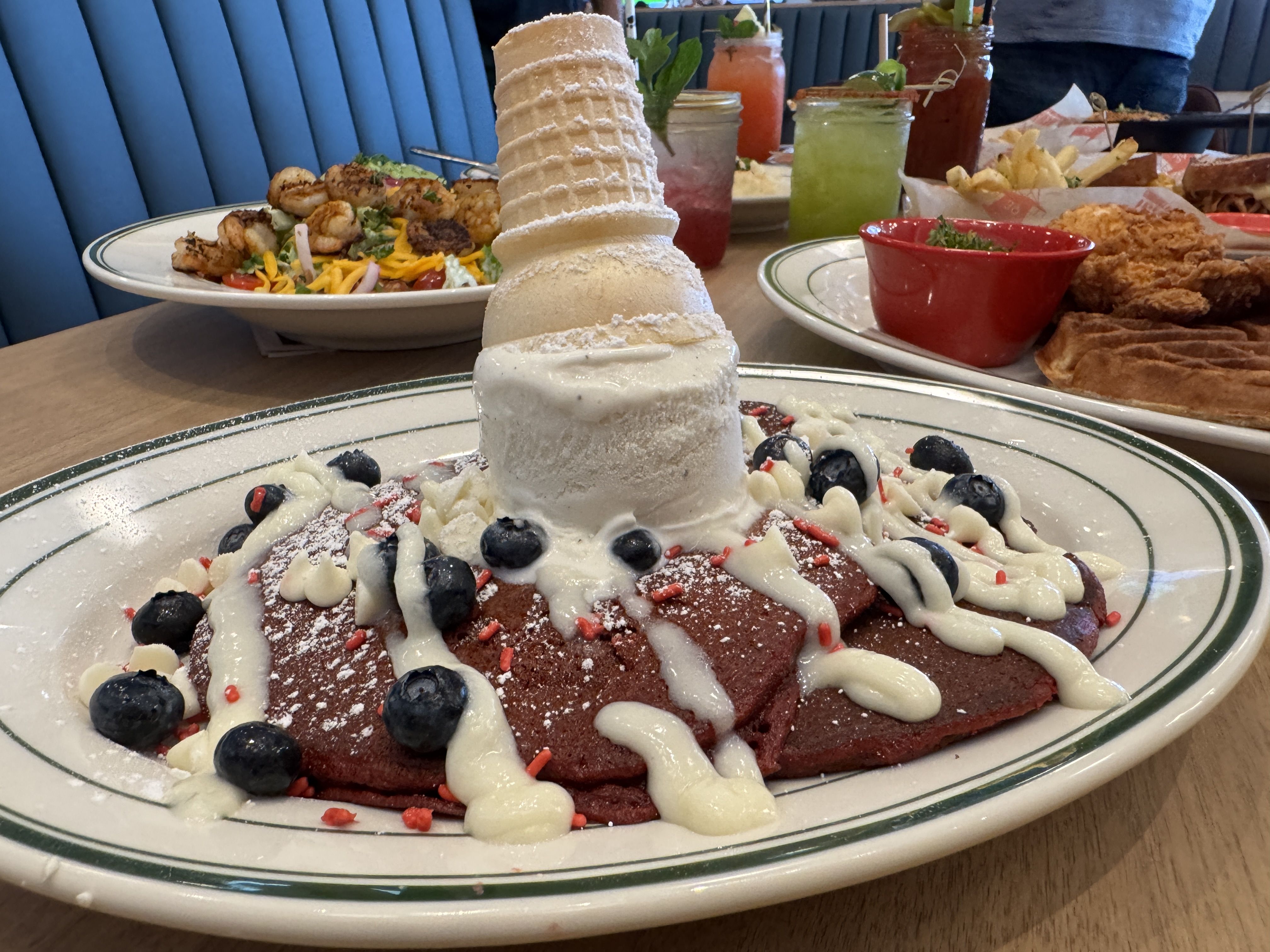 Red velvet pancakes topped with blueberry, white drizzle, powdered sugar, and an upside-down ice cream cone, with other colorful dishes and drinks on a wooden table in a casual restaurant.