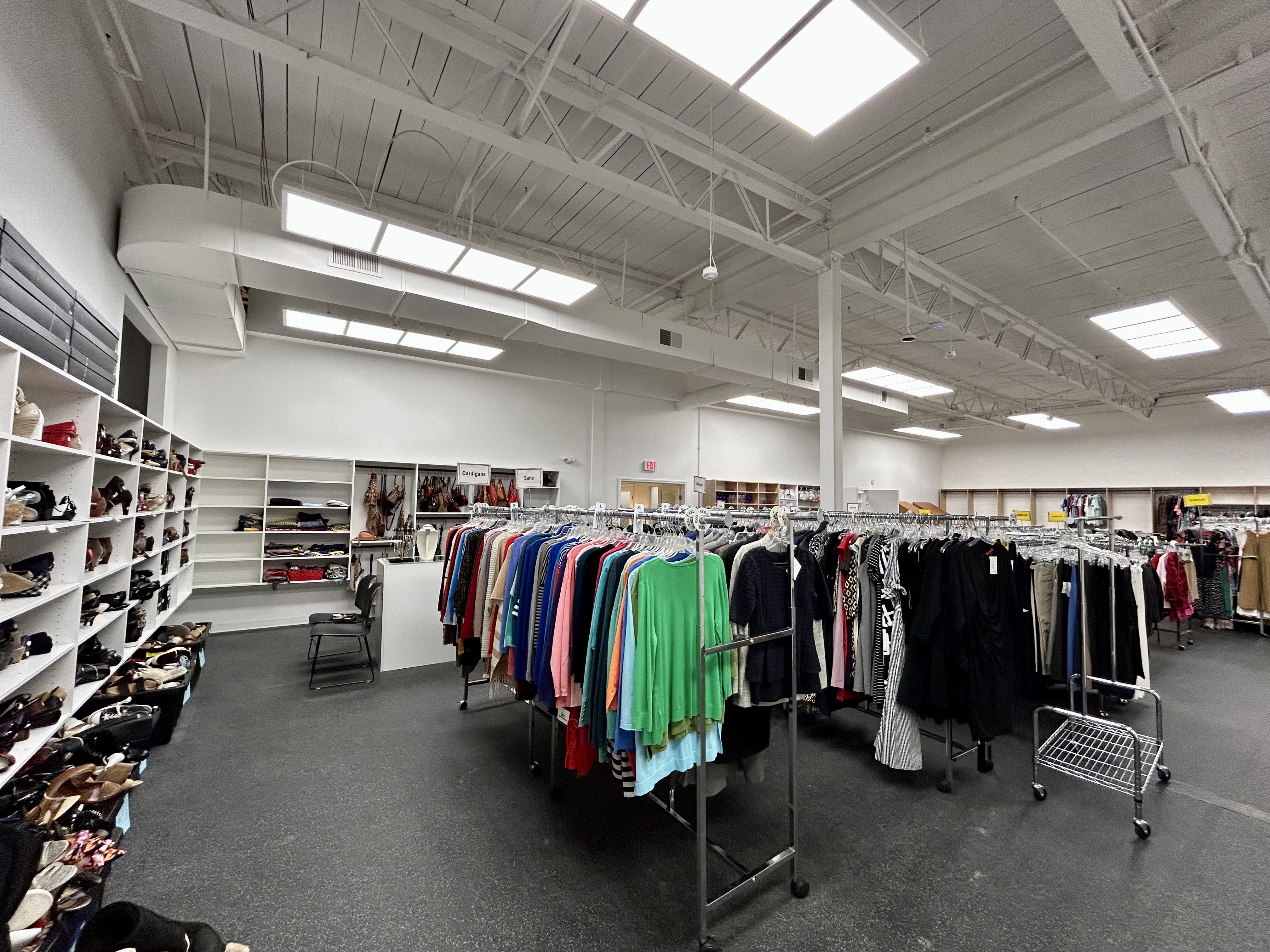 Bright, spacious thrift store interior with racks of colorful clothes, shelves filled with shoes, and white walls under fluorescent ceiling lights.