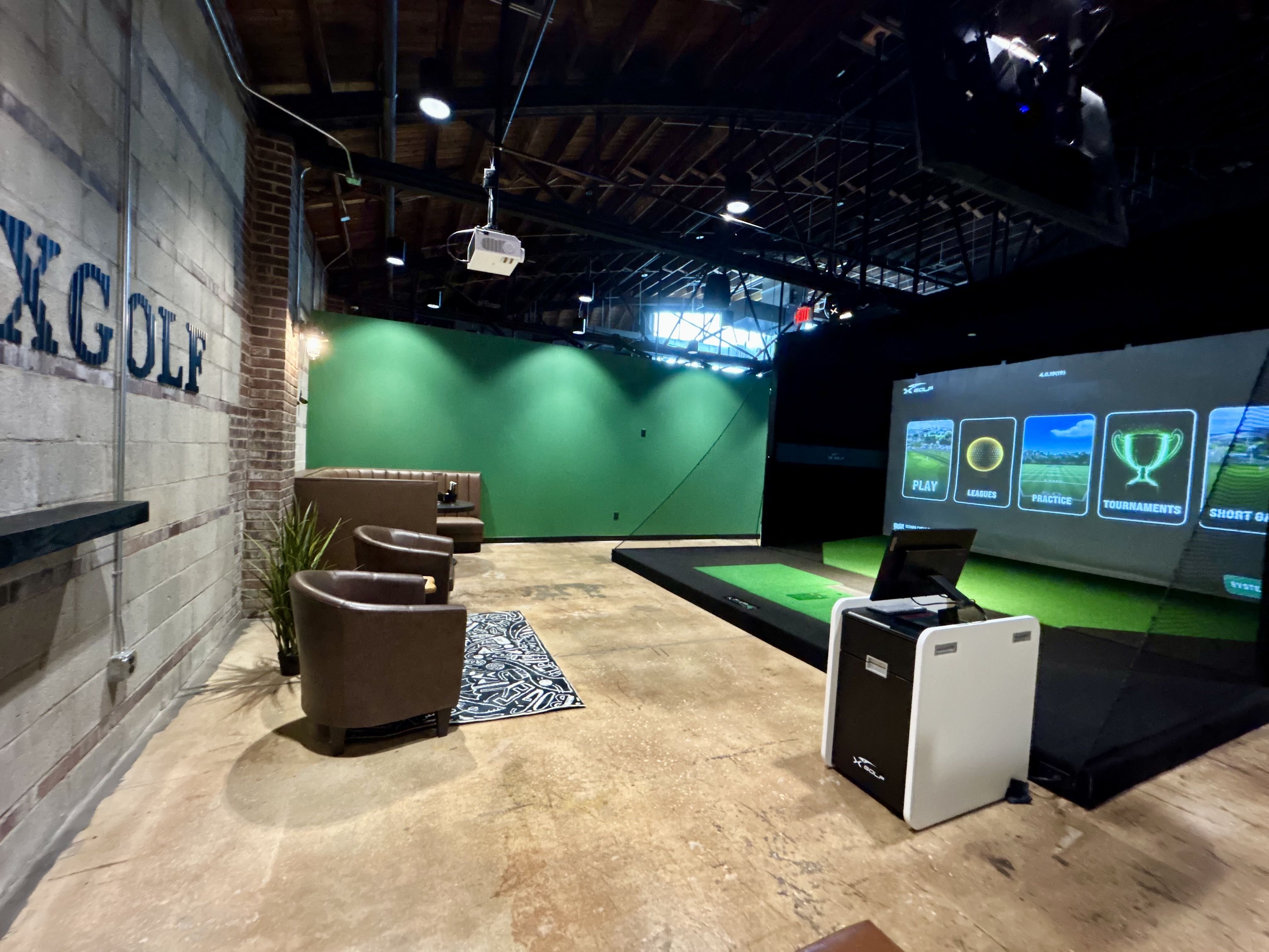 Indoor golf simulation area with a green wall, brown chairs, a projector, and a large screen displaying golf menu options like Play and Tournaments, with industrial ceiling and concrete floor.