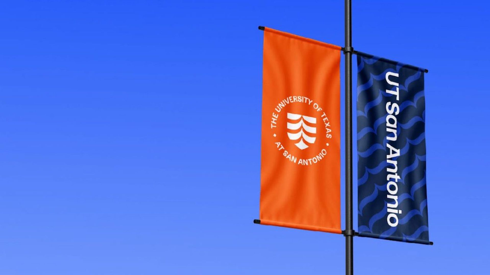 Two vertical banners hanging on a pole against a clear blue sky. Left banner is orange with white text and logo of "The University of Texas at San Antonio." Right banner is dark blue with light blue patterns and white text "UT San Antonio."