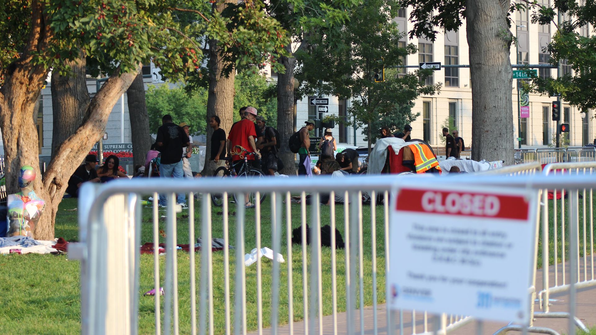 A photo of a park closed sign in the foreground with people experiencing homelessness in the background