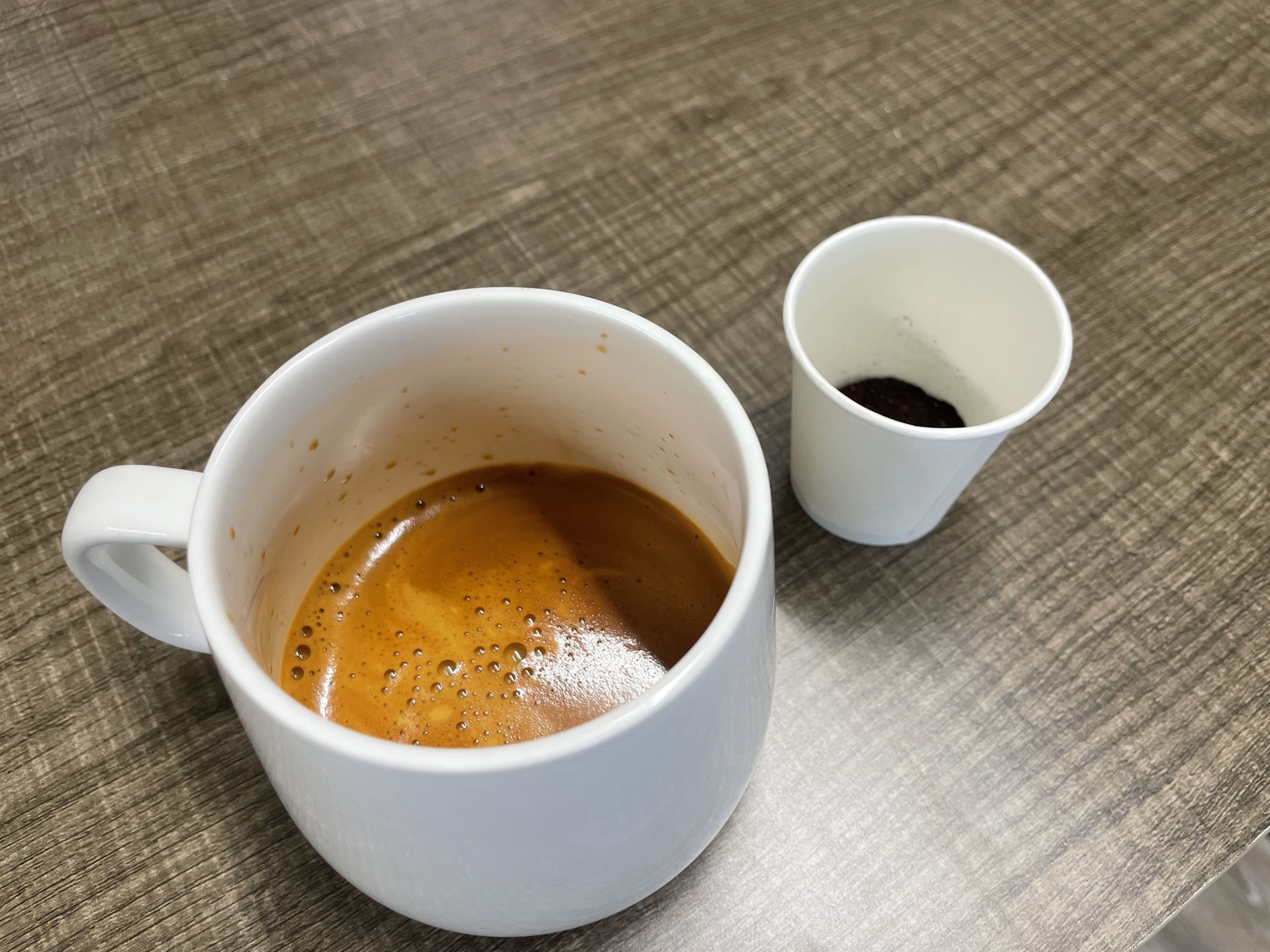 White ceramic cup with freshly brewed espresso on a brown textured table, next to a small white paper cup containing used coffee grounds.