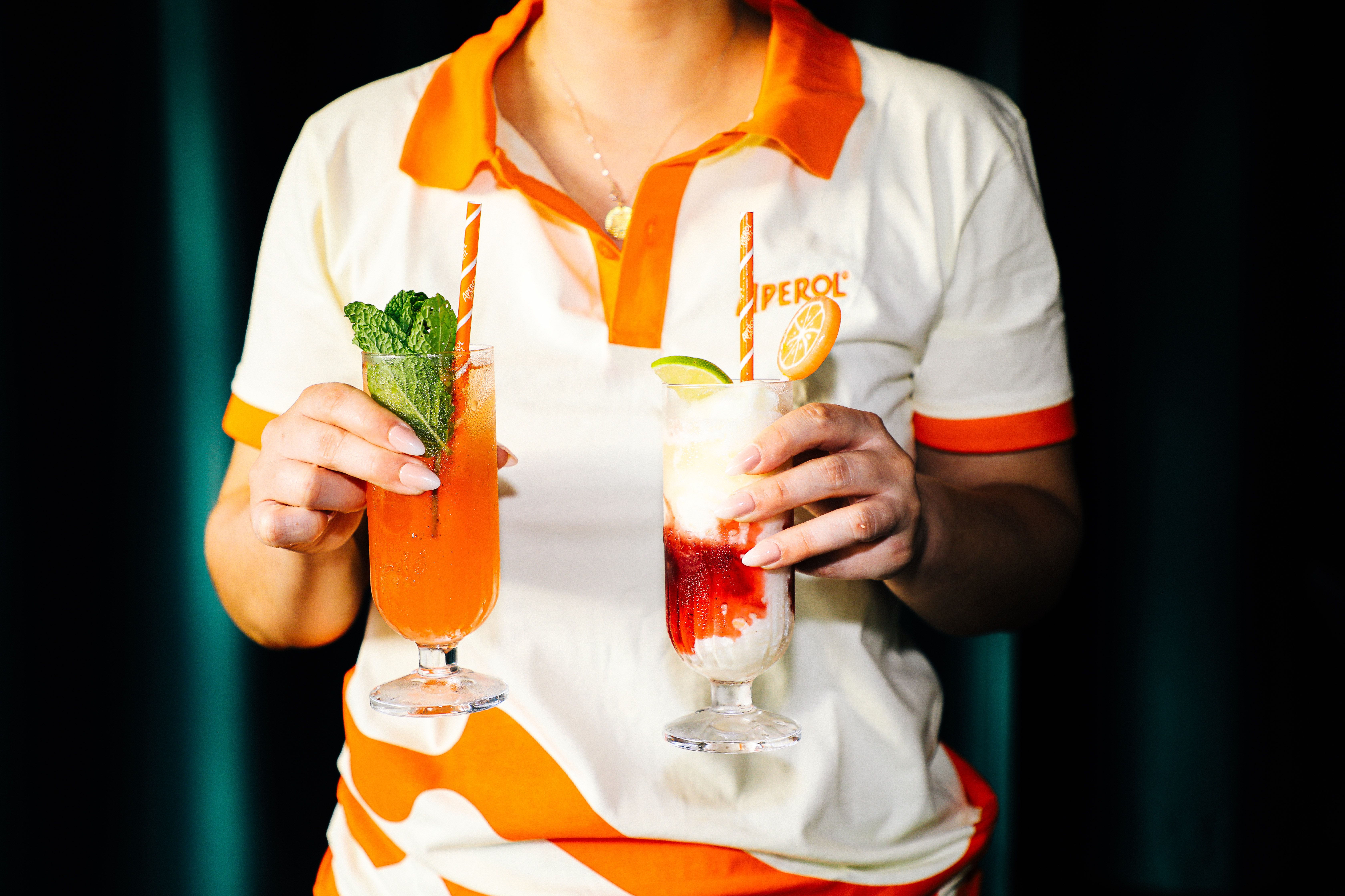A person in a white and orange uniform holds two colorful cocktails in tall glasses: left with bright orange liquid and mint, right with layered red and white, lime and orange garnish, striped straws.