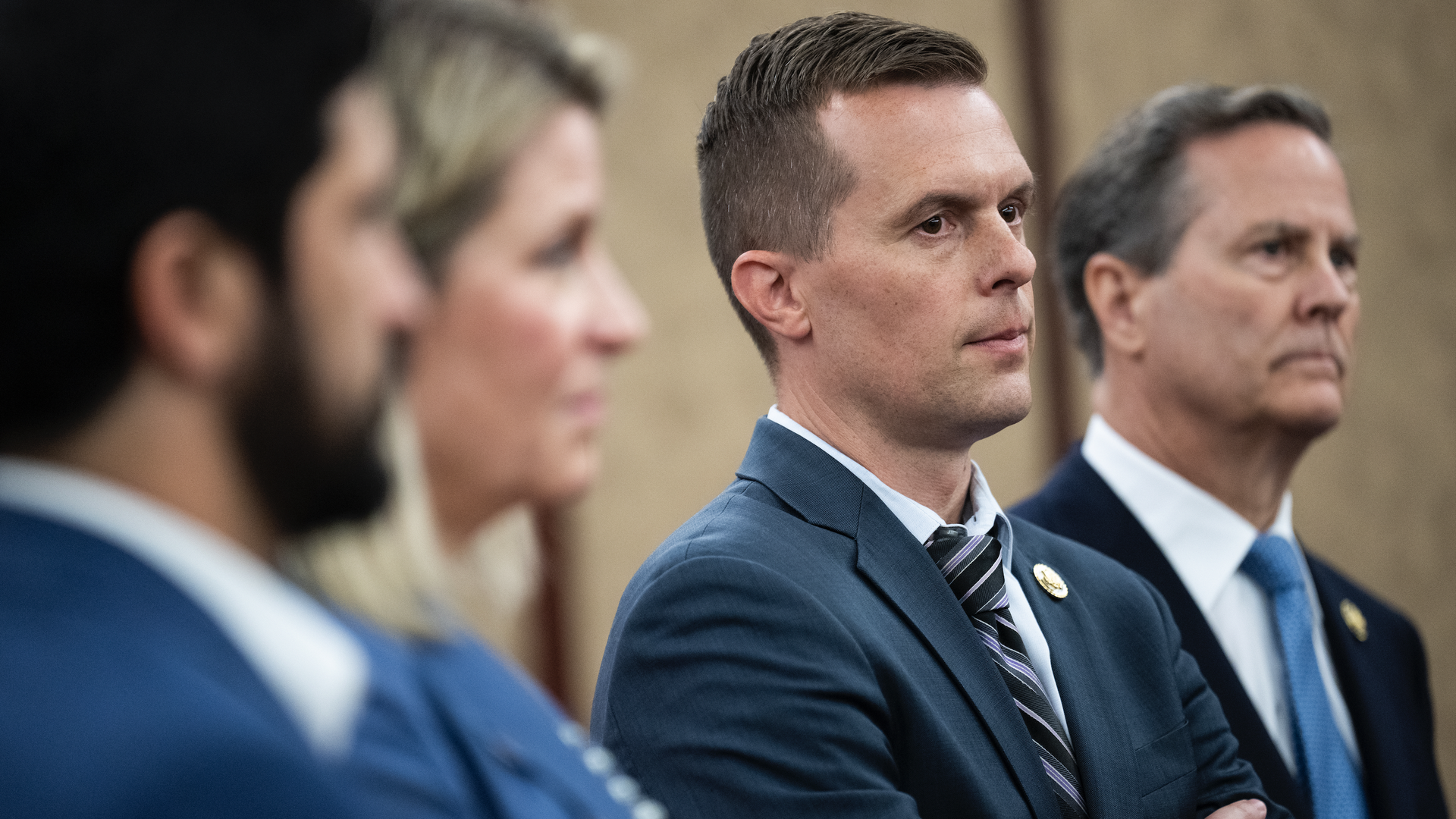 Four people in business attire standing in a line, two men in focus wearing suits and ties, one with arms crossed, neutral background.