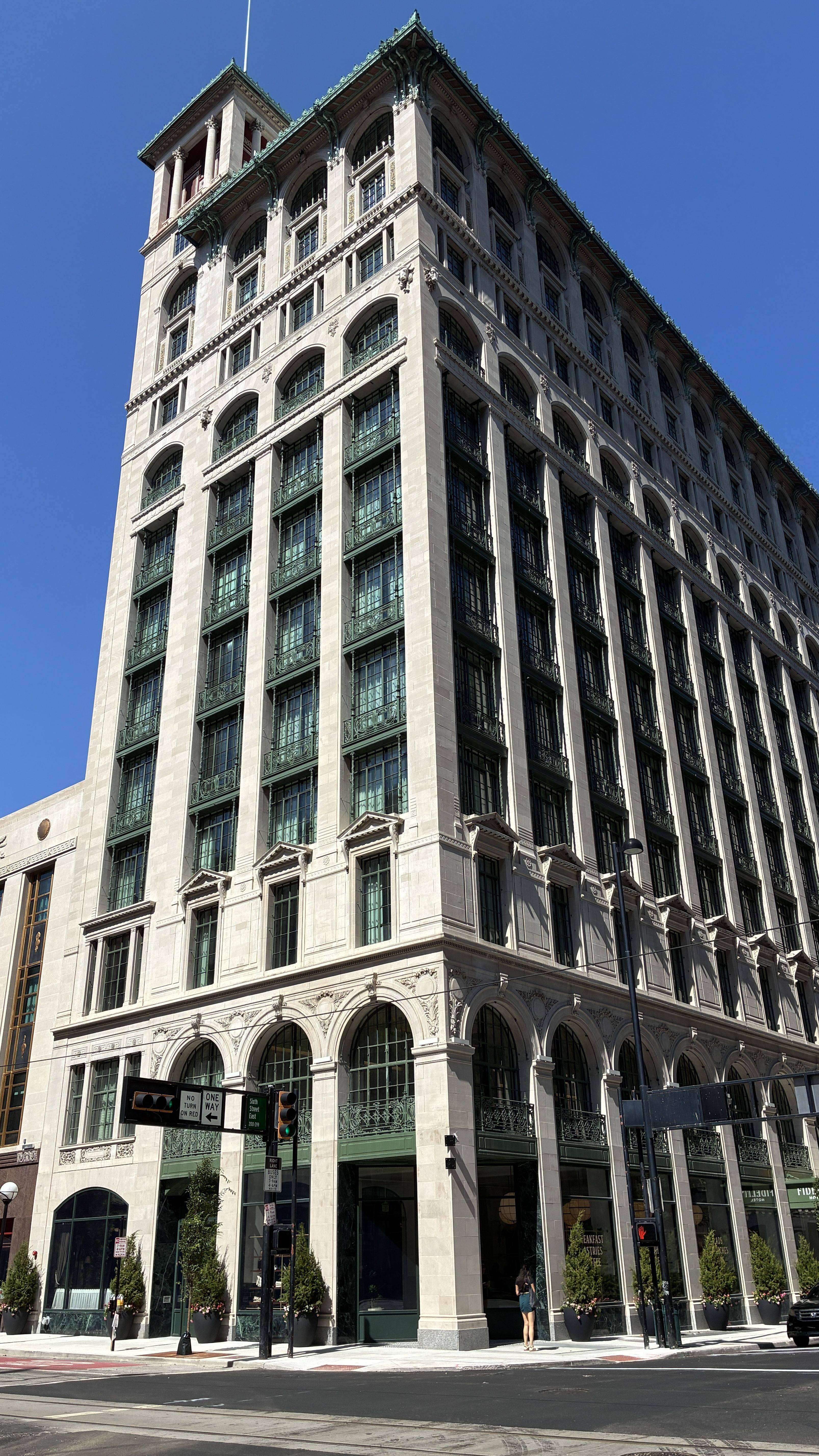 Beige stone skyscraper with arched windows and green wrought-iron balconies, ornate cornices at the top, on a city street with pedestrians and traffic signs under a clear blue sky.