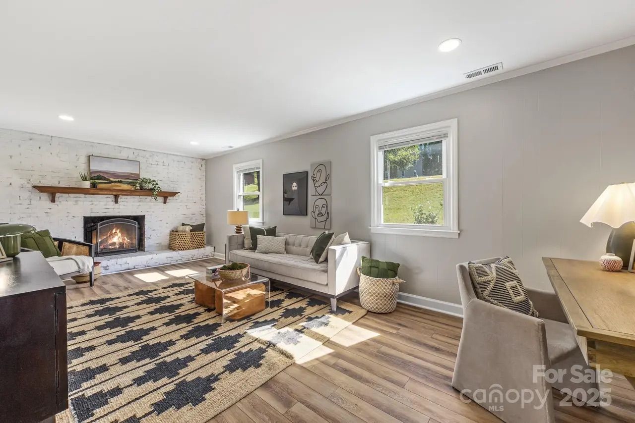 Bright living room with wood floor, beige walls, a white brick fireplace, gray sofa, patterned rug, wooden coffee table, and two windows showing green yard outside.