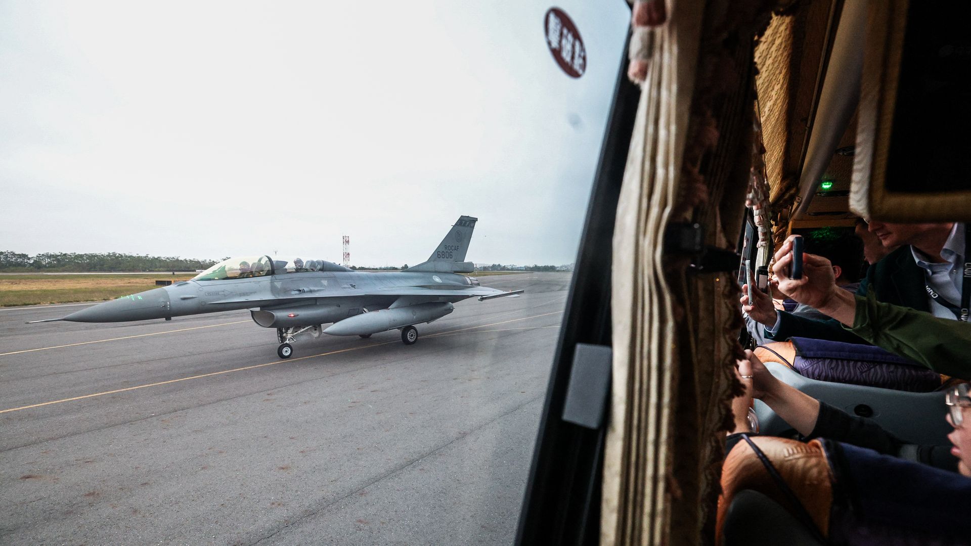 Gray fighter jet with pilots on runway seen through bus window, with passengers inside photographing the aircraft.