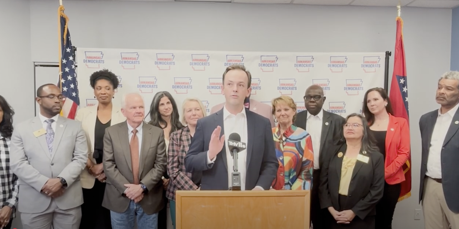 Arkansas state Rep. Andrew Collins (D-Little Rock) stands behind a podium and is flanked by other Democrats.